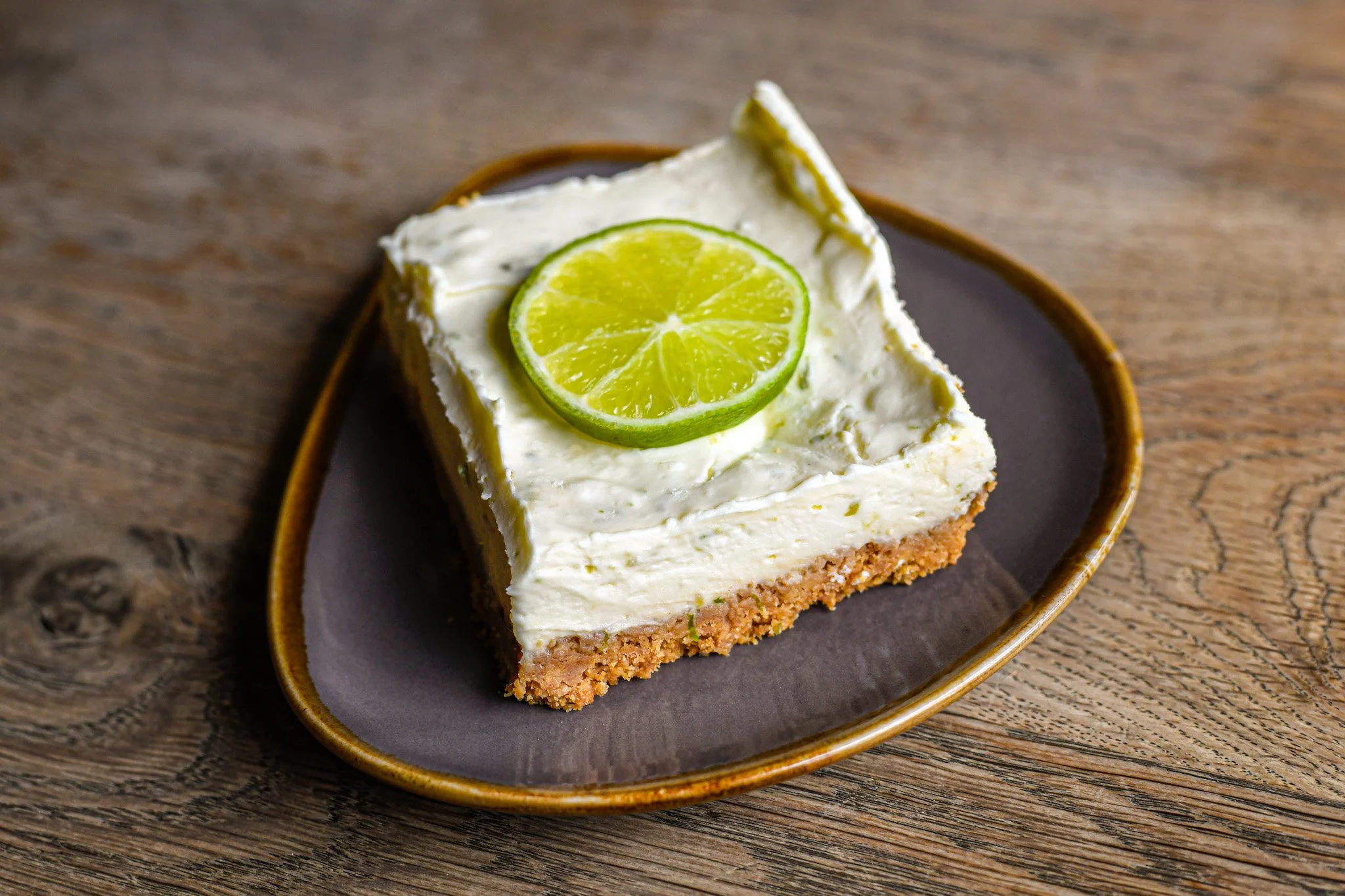 Slice of key lime pie with a lime slice on top, on a dark oval plate on a wooden table.