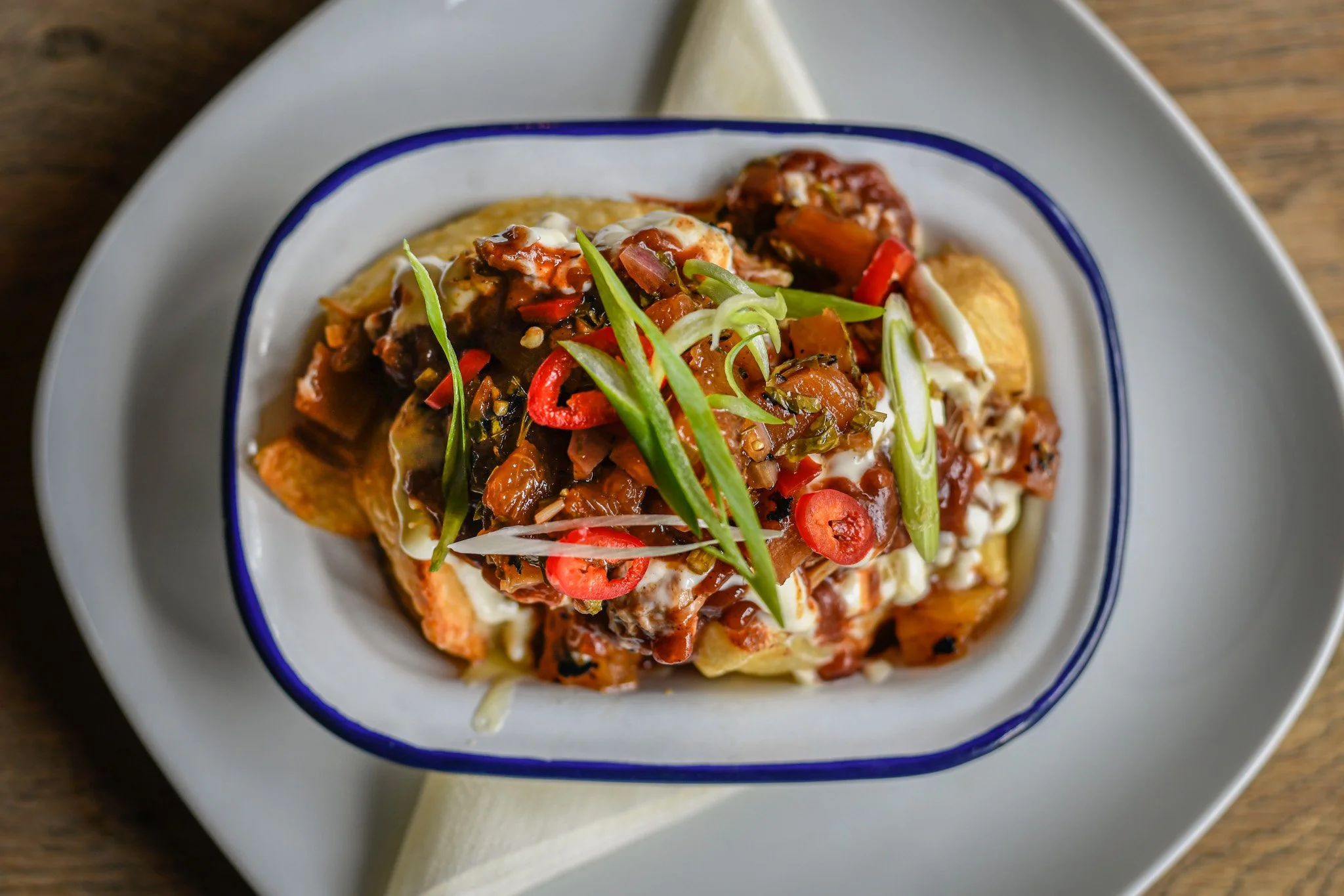 A bowl of loaded nachos topped with sliced green onions, red chili peppers, chopped vegetables, and sauce, served on a white plate with a paper napkin.
