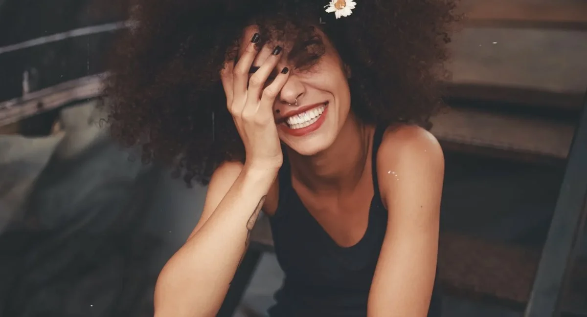 Woman with curly hair and tattoos laughing with one hand covering part of her face while sitting on wooden stairs, symbolizing emotional release and healing through burnout recovery in Salt Lake City.