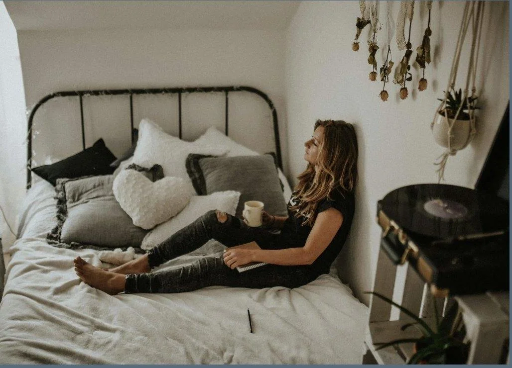 Woman sitting on a bed holding a mug with eyes closed and a peaceful expression in a cozy bedroom with decorative pillows and dried flowers, symbolizing emotional regulation and somatic therapy in Salt Lake City.