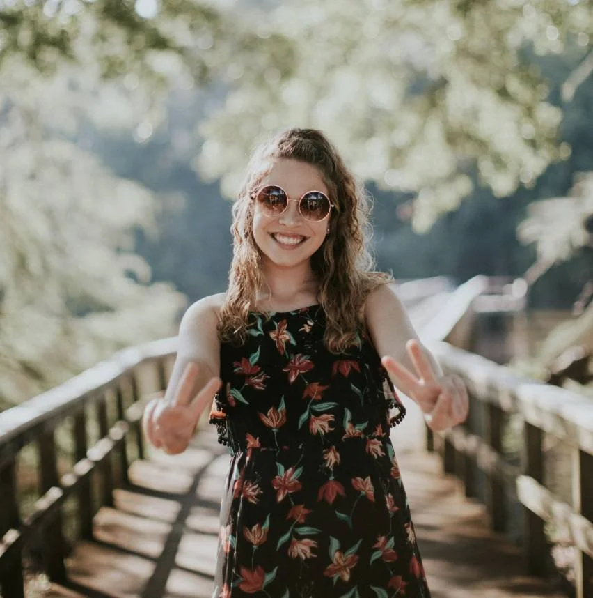 Woman with curly hair in sunglasses and floral dress smiling with peace signs on a wooden bridge, symbolizing burnout recovery in Salt Lake City.