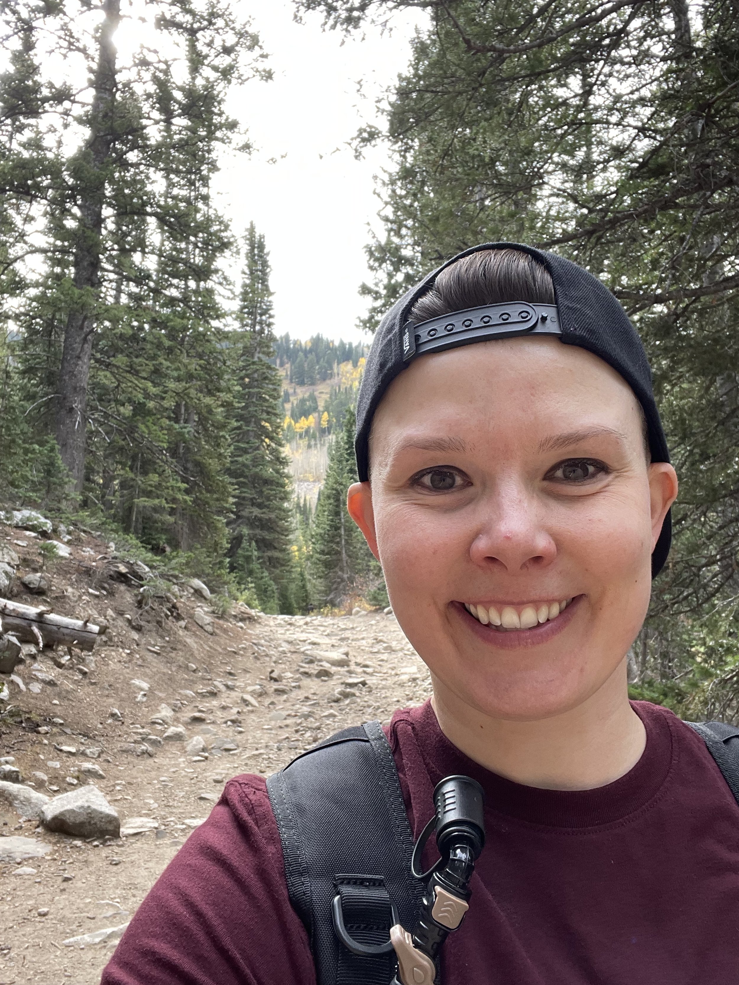 Smiling woman with a backpack taking a selfie on a forest trail.