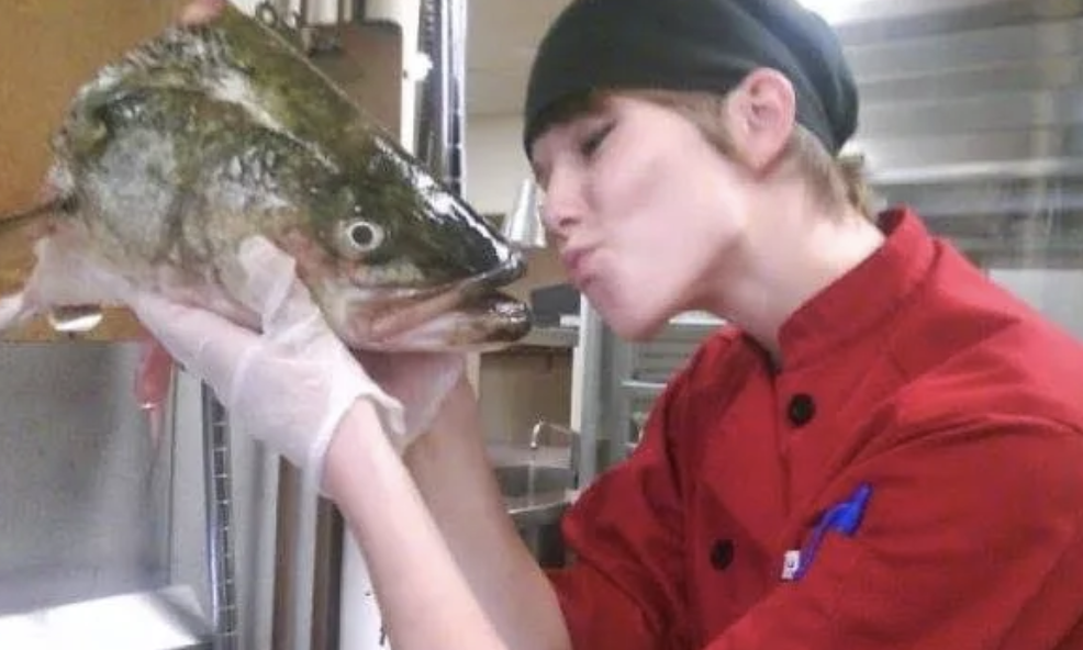 A young person in a chef's coat and hat is holding a large fish close to their face, preparing to kiss it.
