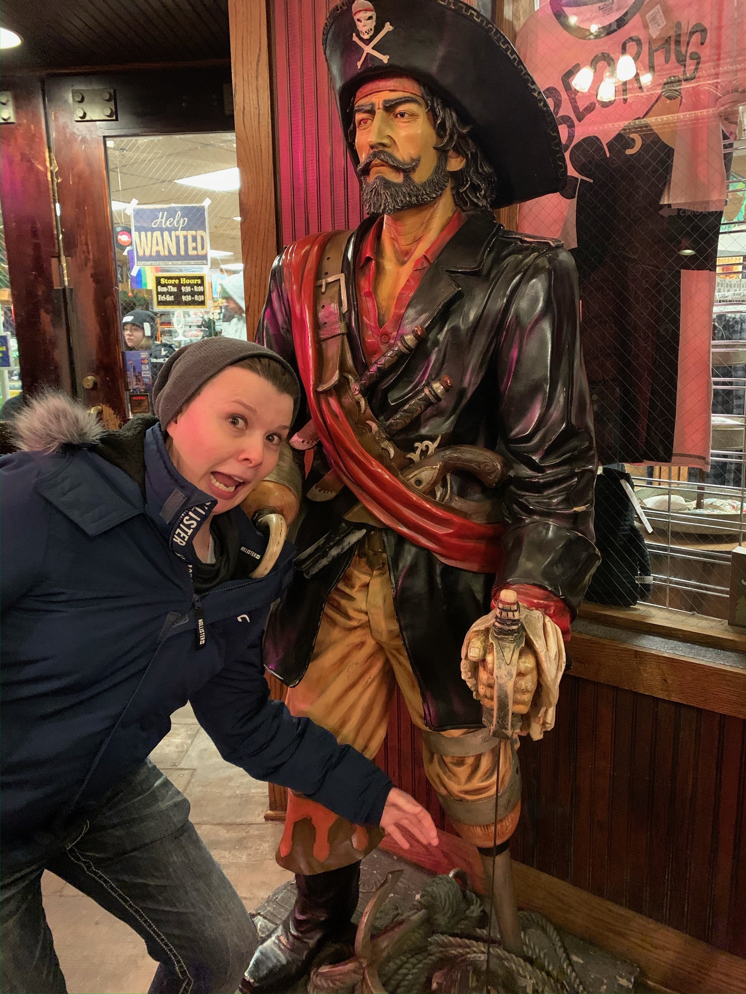 A woman with a surprised expression crouching next to a large pirate statue holding a sword. The pirate is dressed in traditional attire with a large black hat, a red sash, and a leather coat, and has a beard and mustache. The setting appears to be i