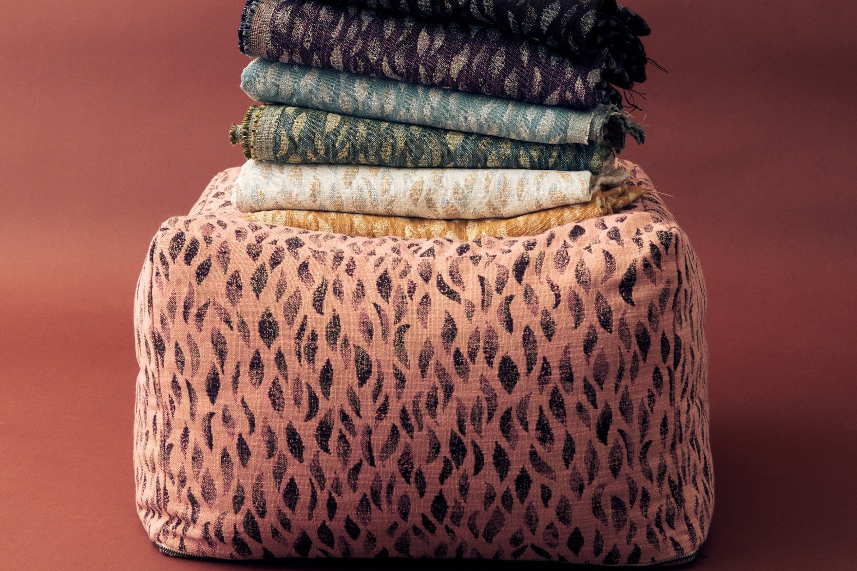 An ottoman upholstered in Petals Woven Rose/Purple, topped with a stack of other colorways