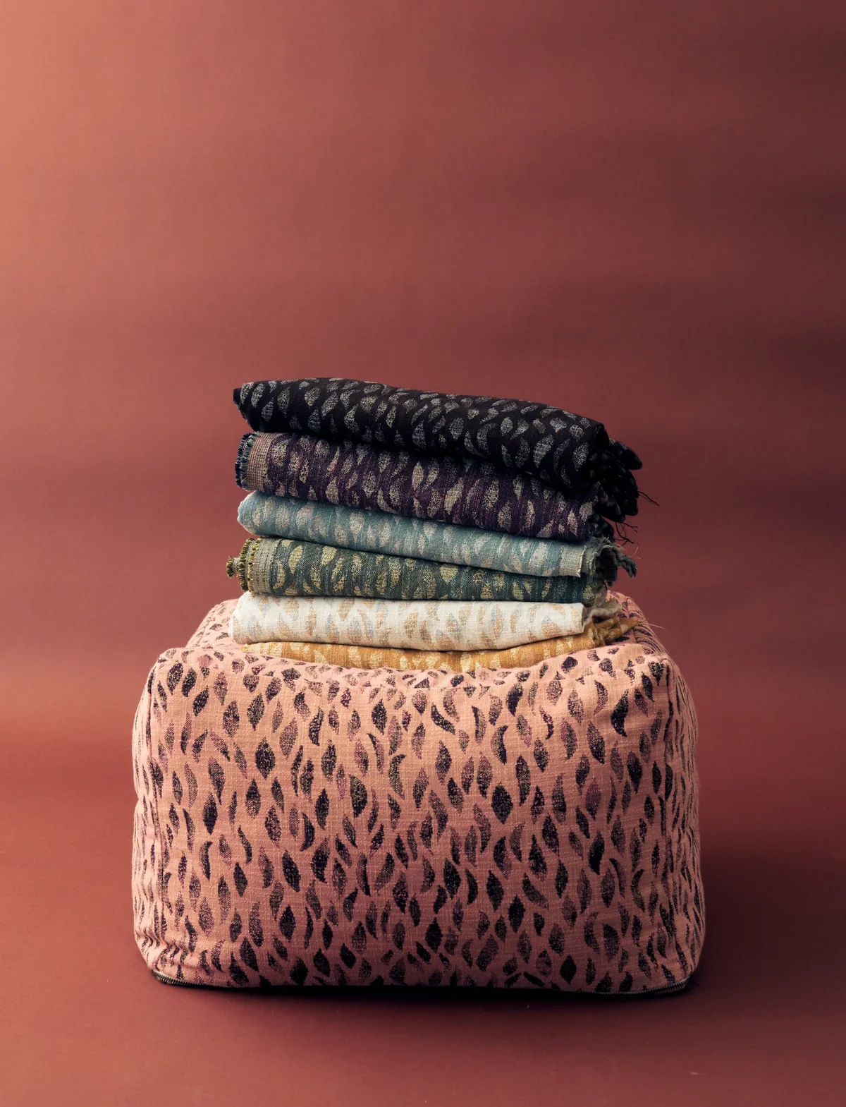 A stack of Rebecca Atwood Petals woven fabric in six colorways resting on a pink Petals upholstered pouf