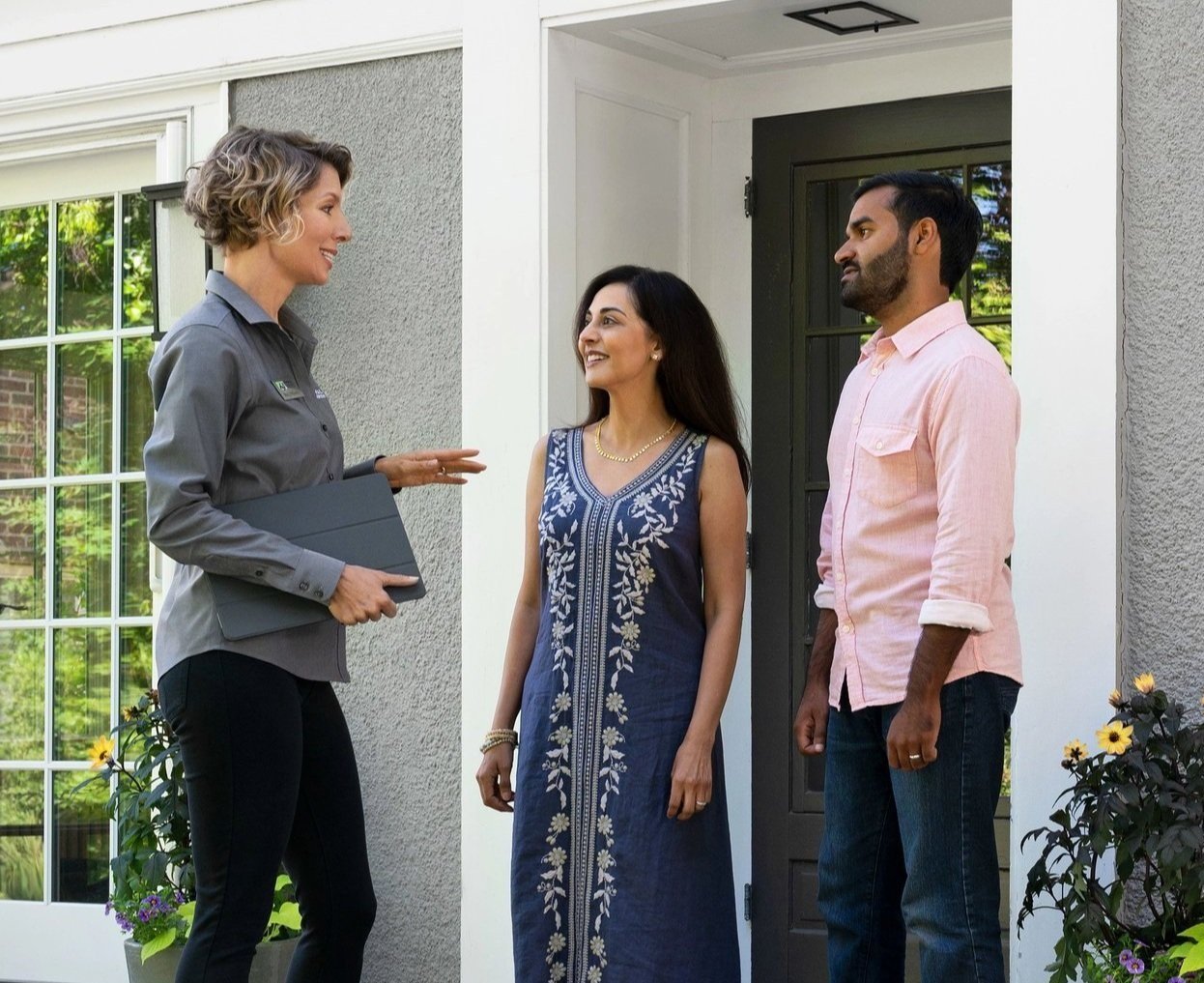 Design consultant meeting with couple outside a home: a woman on the left wearing a grey shirt and holding a tablet, speaking to a woman and a man standing in a doorway.