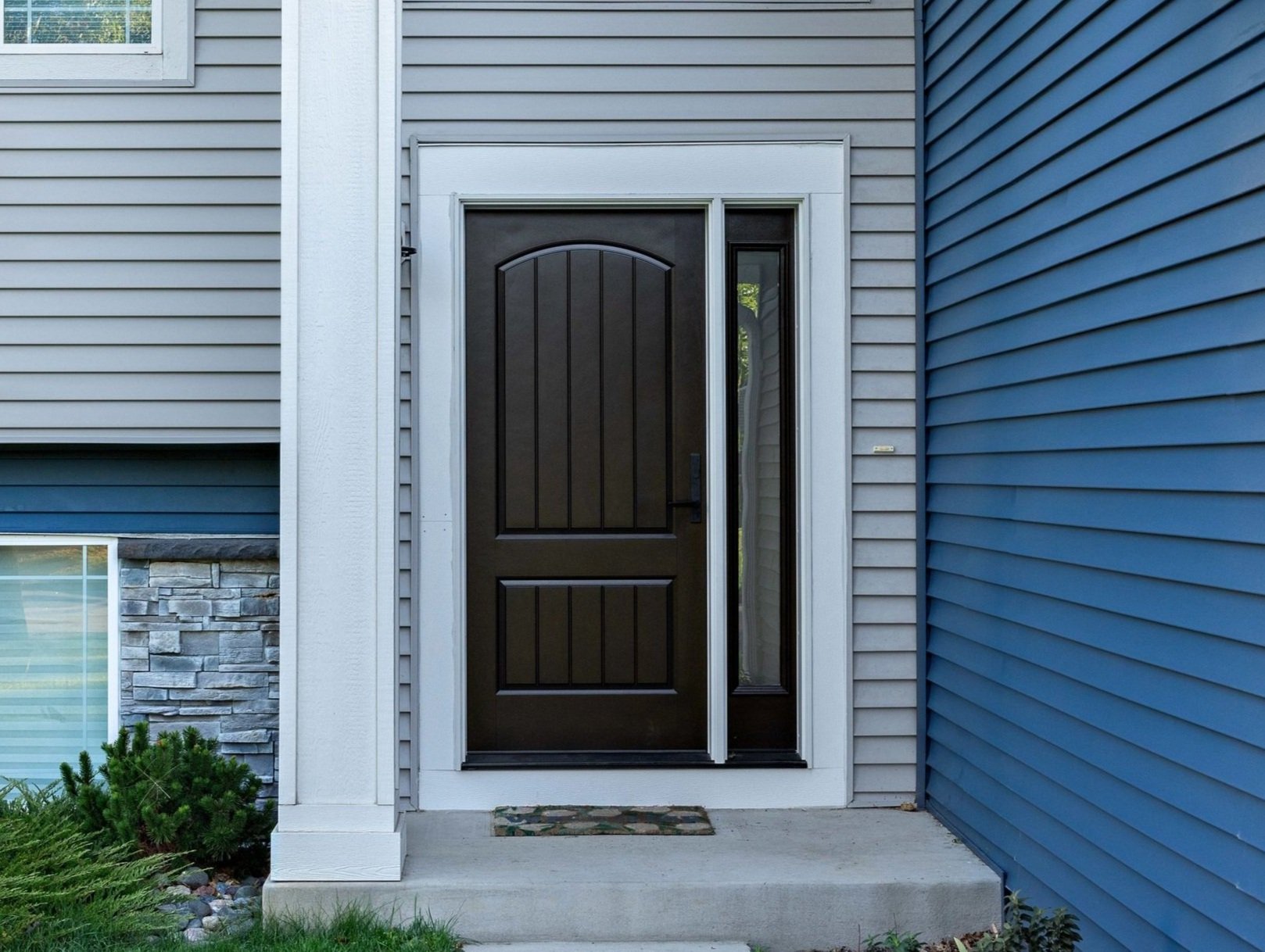 Front of a house showing a dark wooden entry door with a sidelight window, gray and blue vinyl siding, a welcome mat, and landscaping with shrubs.