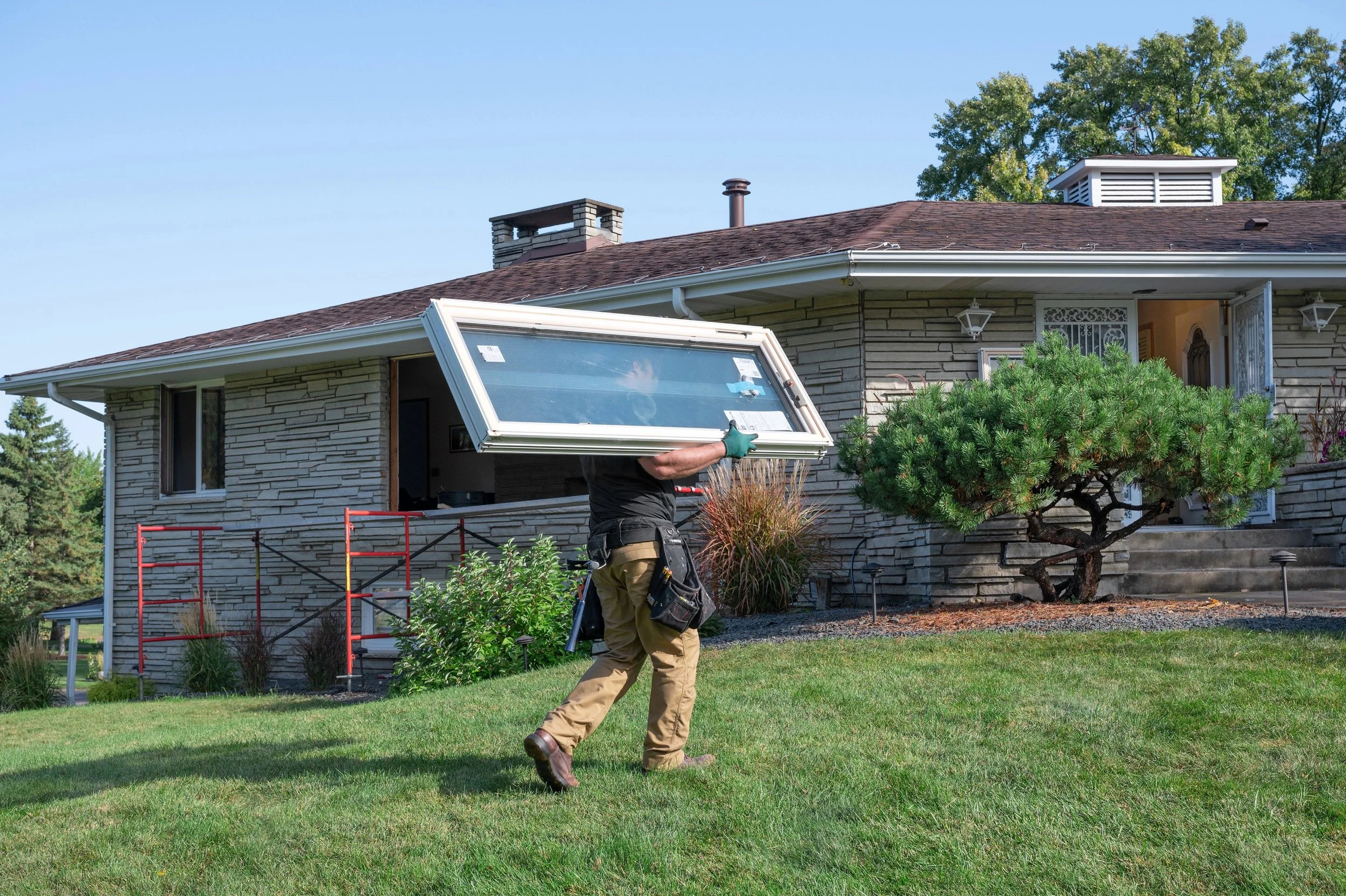 A Renewal by Andersen professional installer is carrying a large replacement window frame in front of a stone house.