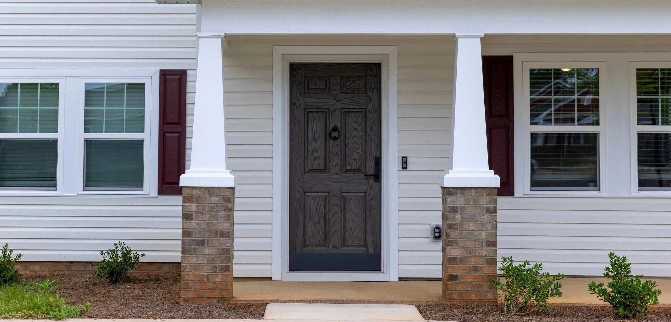 The house has a dark wooden front door, white siding, brick pillars, and horizontal windows with shutters.