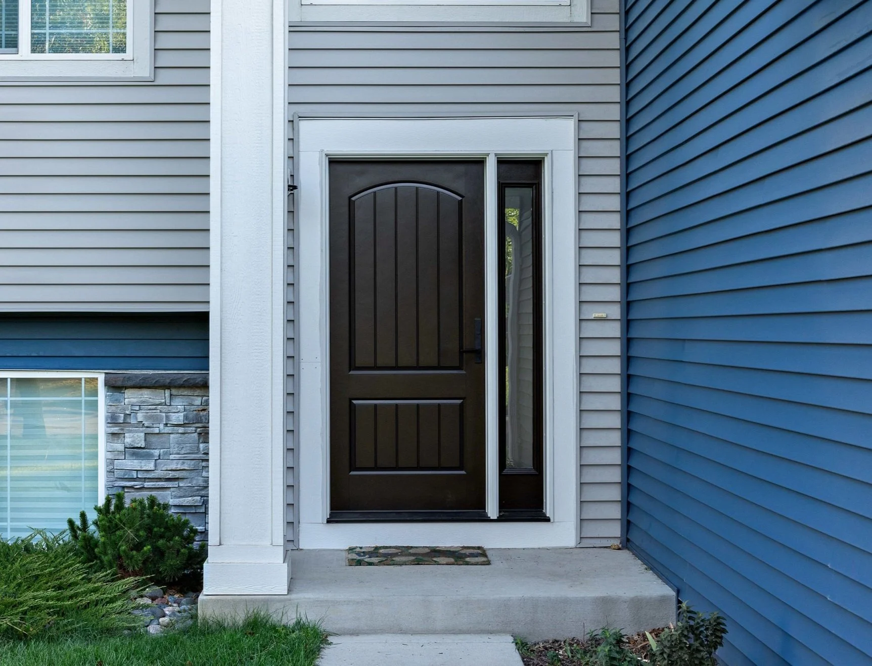 Front door of a house with dark brown panel with a sidelight window on the right side, surrounded by light gray siding and decorative stone accents.