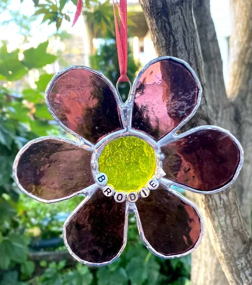 A stained glass flower ornament with pink petals and a yellow center hanging from a pink ribbon on a tree branch in a garden.
