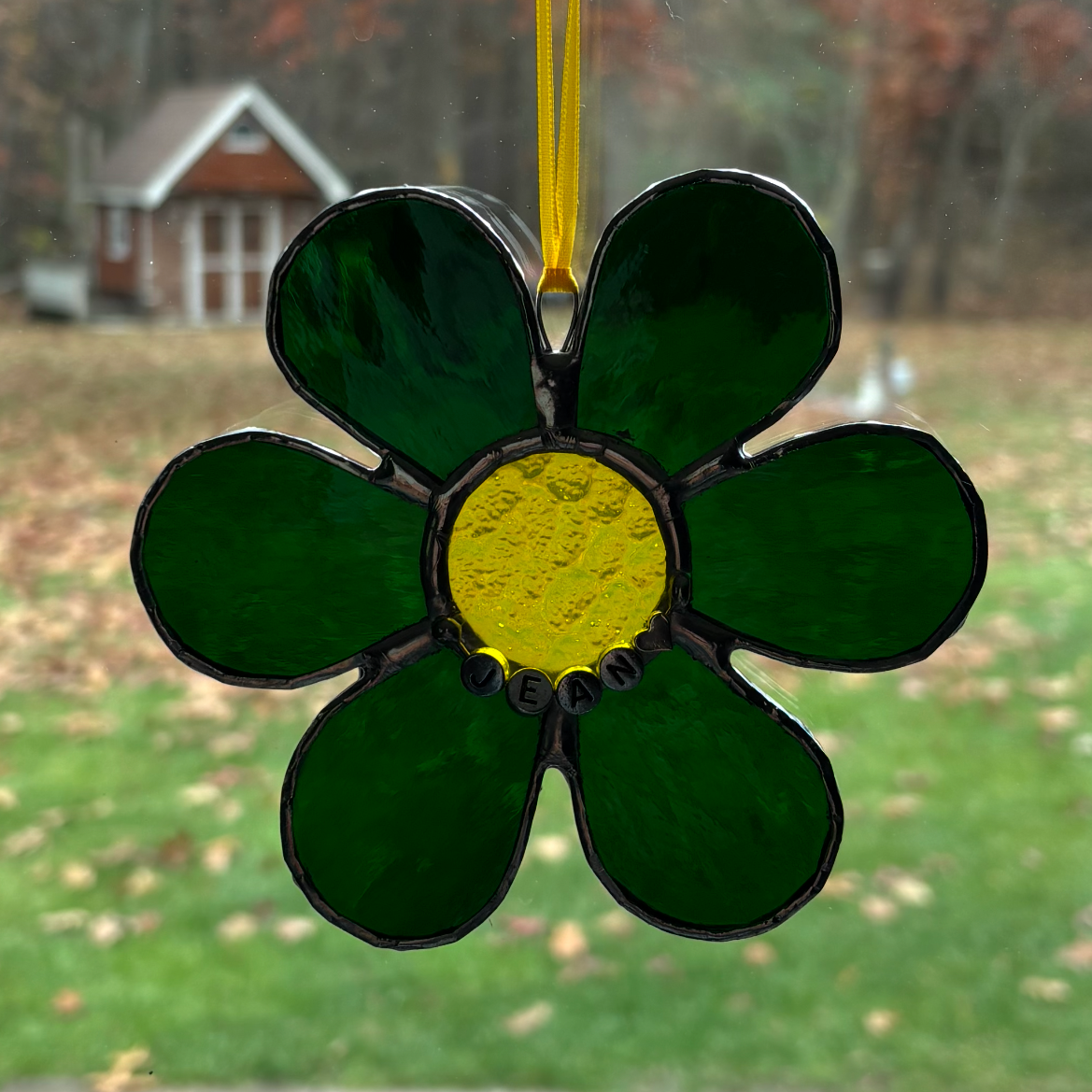 Stained glass ornament in the shape of a flower with green petals and a yellow center, hanging in front of a window with a blurred outdoor view.