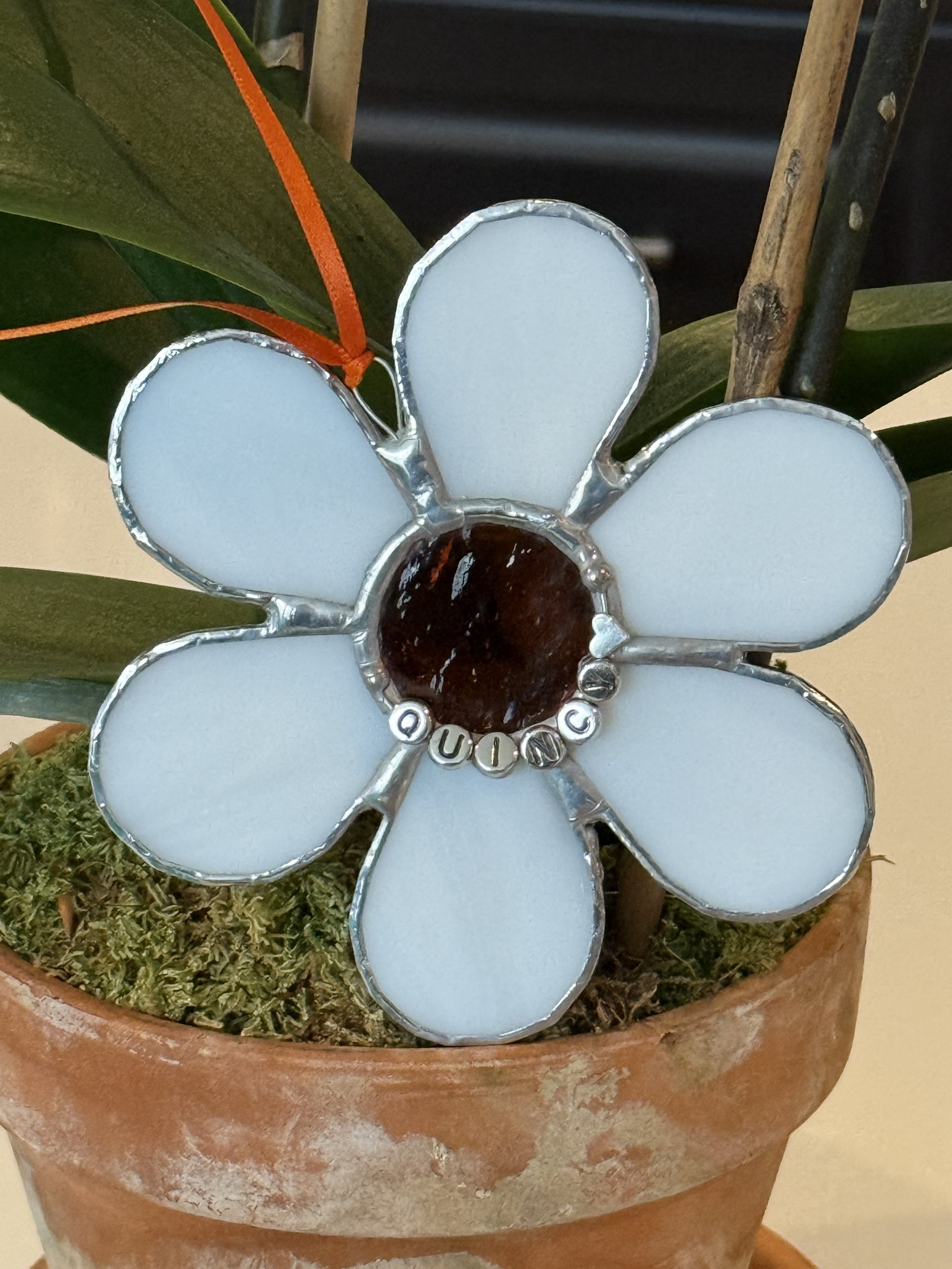 A flower-shaped ornament with white petals and a dark center, featuring the word 'QUINCY' spelled out in small white beads, hanging from an orange ribbon on a potted plant.