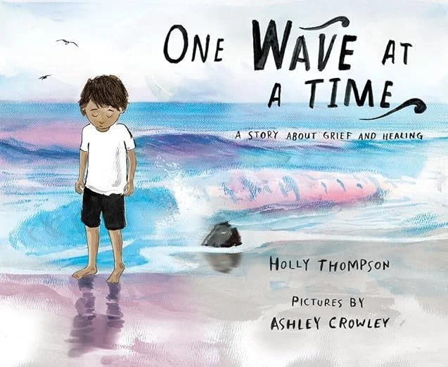 A children's book cover titled 'One Wave at a Time: A Story About Grief and Healing' by Holly Thompson, illustrated by Ashley Crowley. The cover features a young boy standing barefoot on the beach near the water, looking down with a somber expression