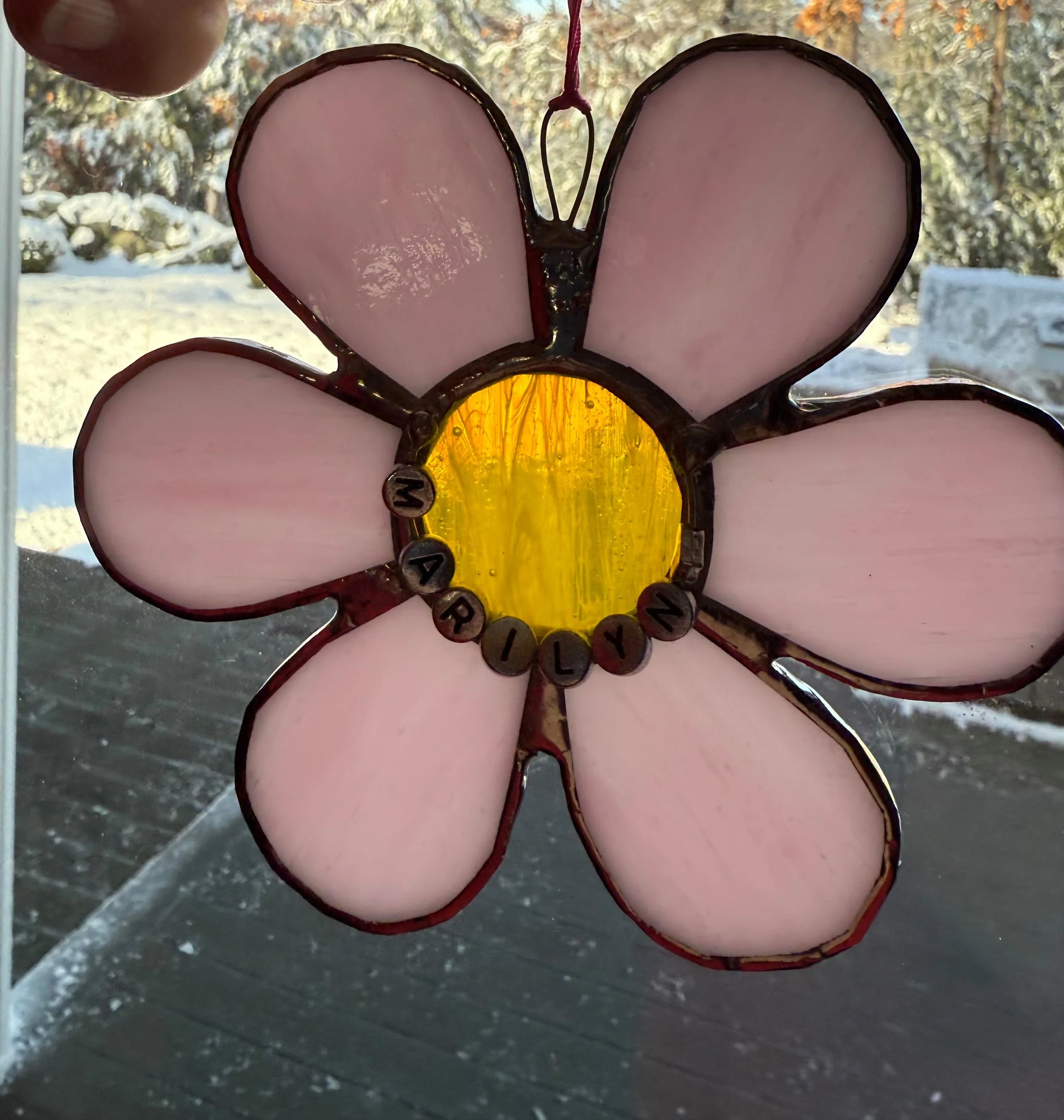 A stained glass flower with pink petals and a yellow center, spelling out the word 'MARILYN' with letter beads.