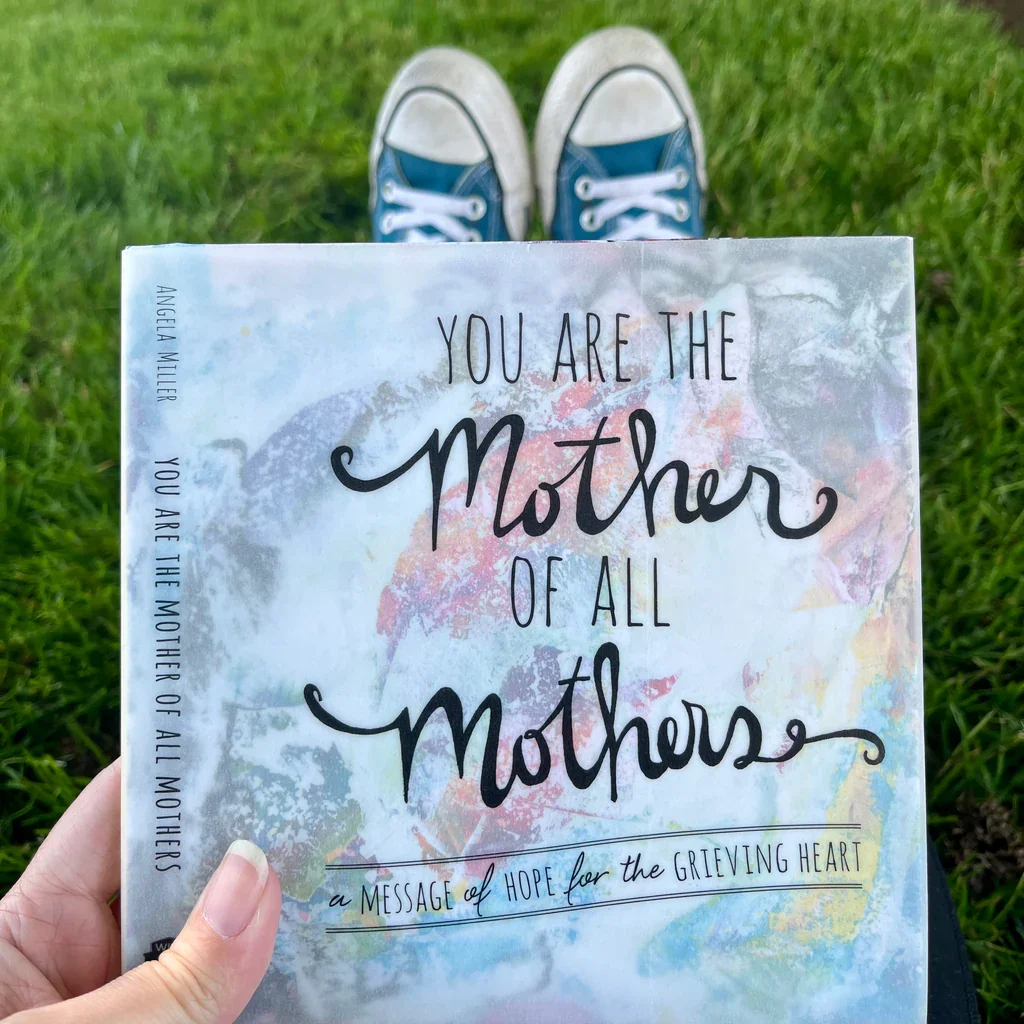 Hand holding a book titled "You Are the Mother of All Mothers" with a background of grass and feet in blue sneakers.