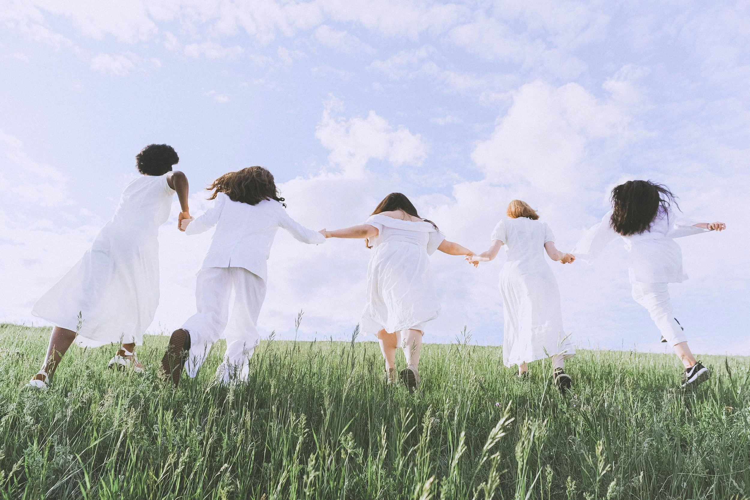 Five women in white dresses running hand-in-hand across a grassy field under a partly cloudy sky.