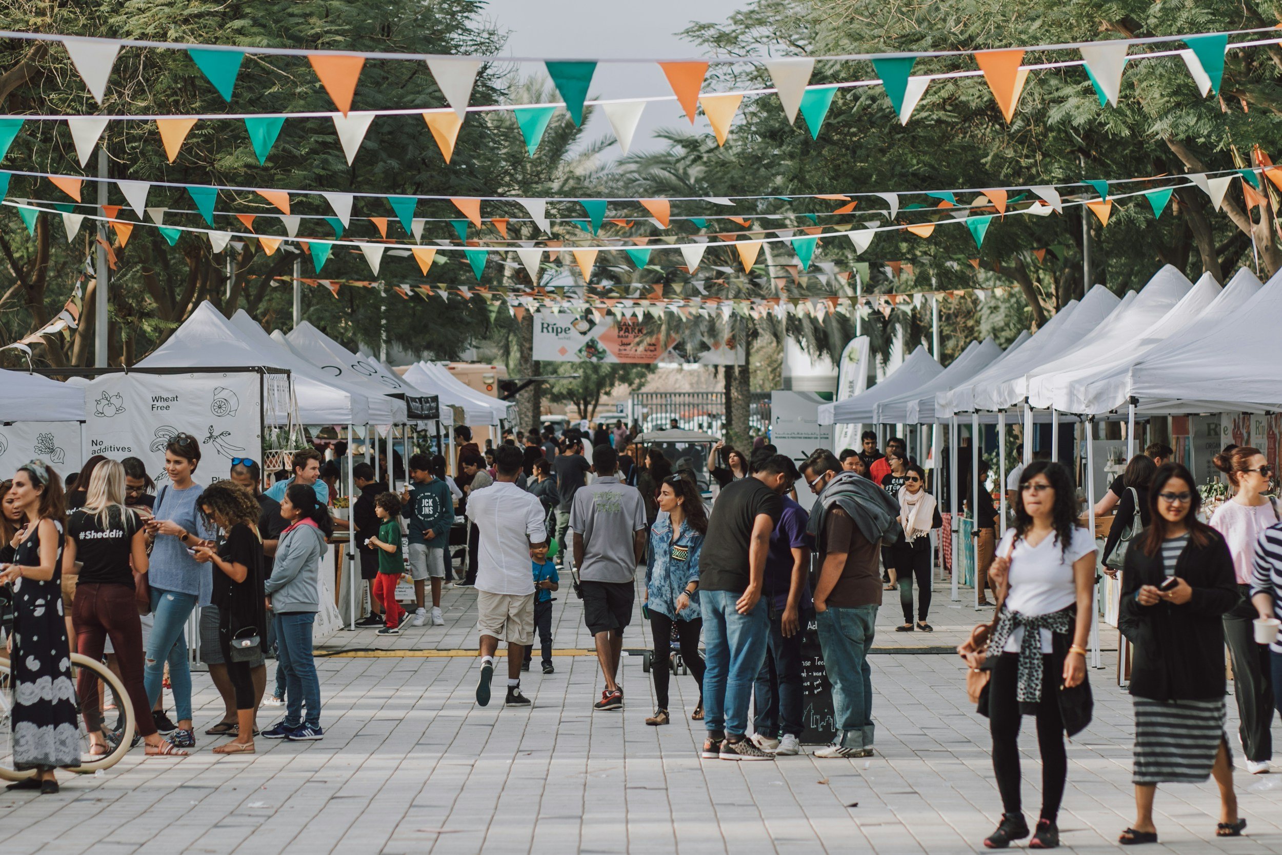 People walking and browsing at an outdoor market or fair with vendor tents and colorful triangular flags hanging overhead.