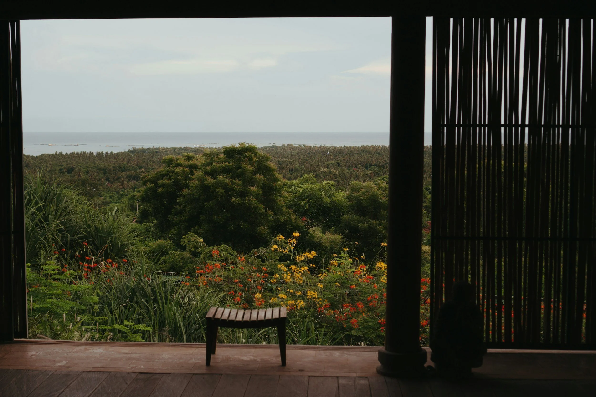 View of lush green trees and colorful flowers through an open window with a wooden stool and a small statue in the foreground, and the ocean visible in the distance under a cloudy sky