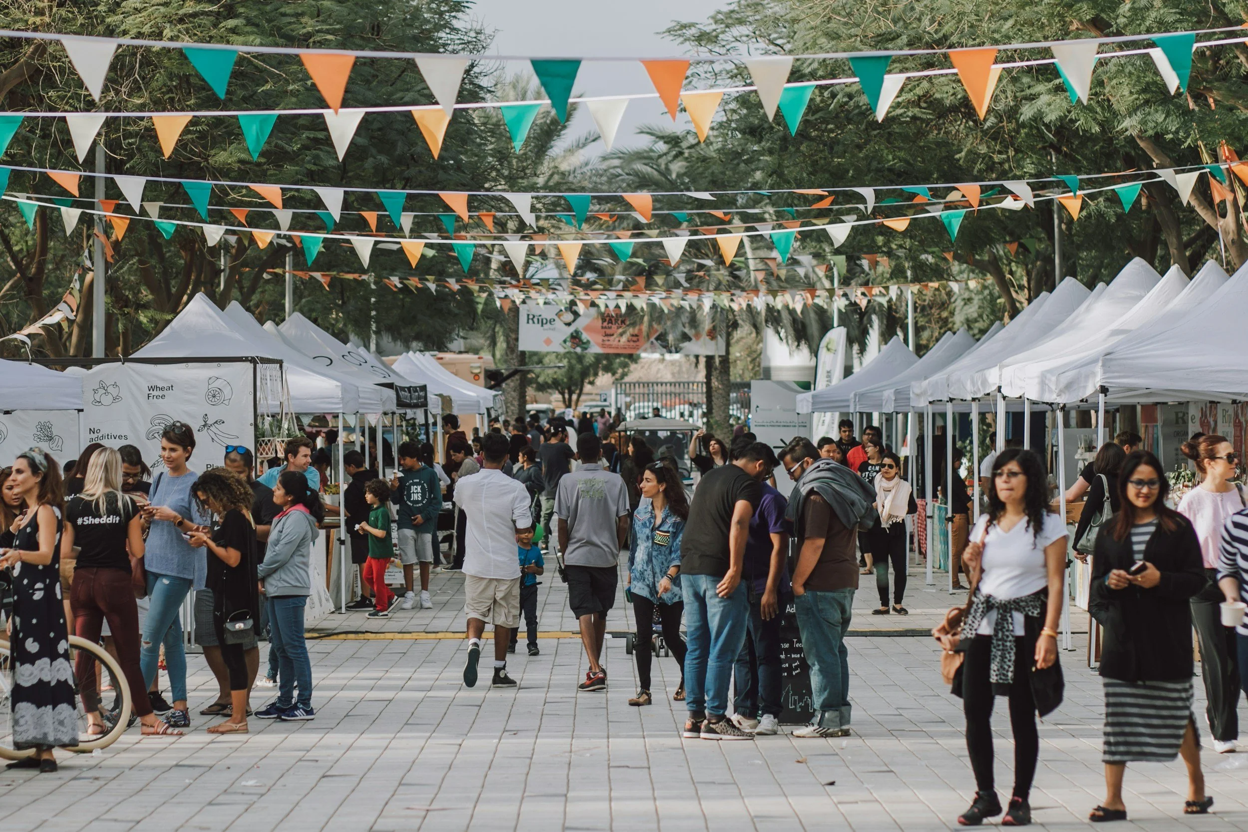 A lively outdoor market or fair with white tents and colorful triangular flags overhead, with many people walking, shopping, and socializing.