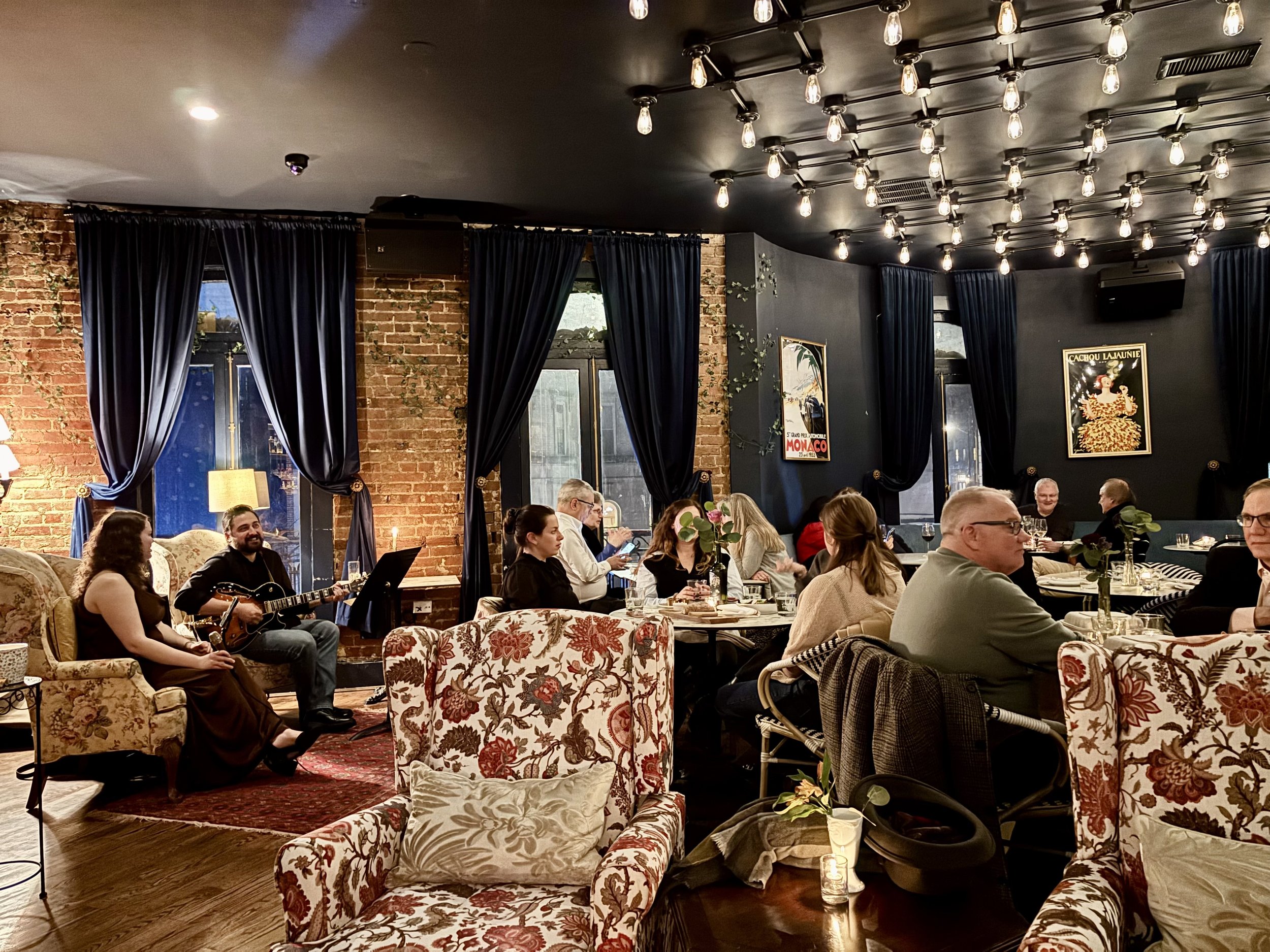 A cozy restaurant interior featuring exposed brick walls, large dark blue curtains, framed artwork, and a mix of floral and plain upholstered seating. There is live music being performed by a guitarist and a singer among the diners.