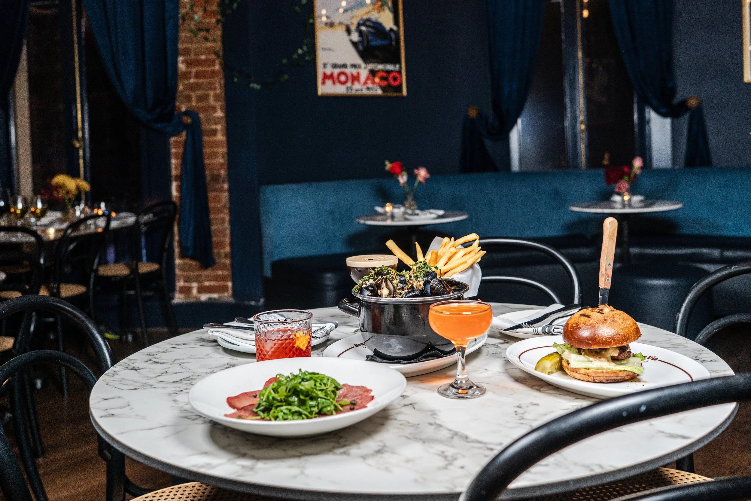A dining table set with various dishes including a burger with lettuce and pickles, a plate of pasta with sauce, and a bowl of mussels. There are also glasses of drinks on the table. The background shows a cozy restaurant interior with blue walls, curtains, and some wall art.