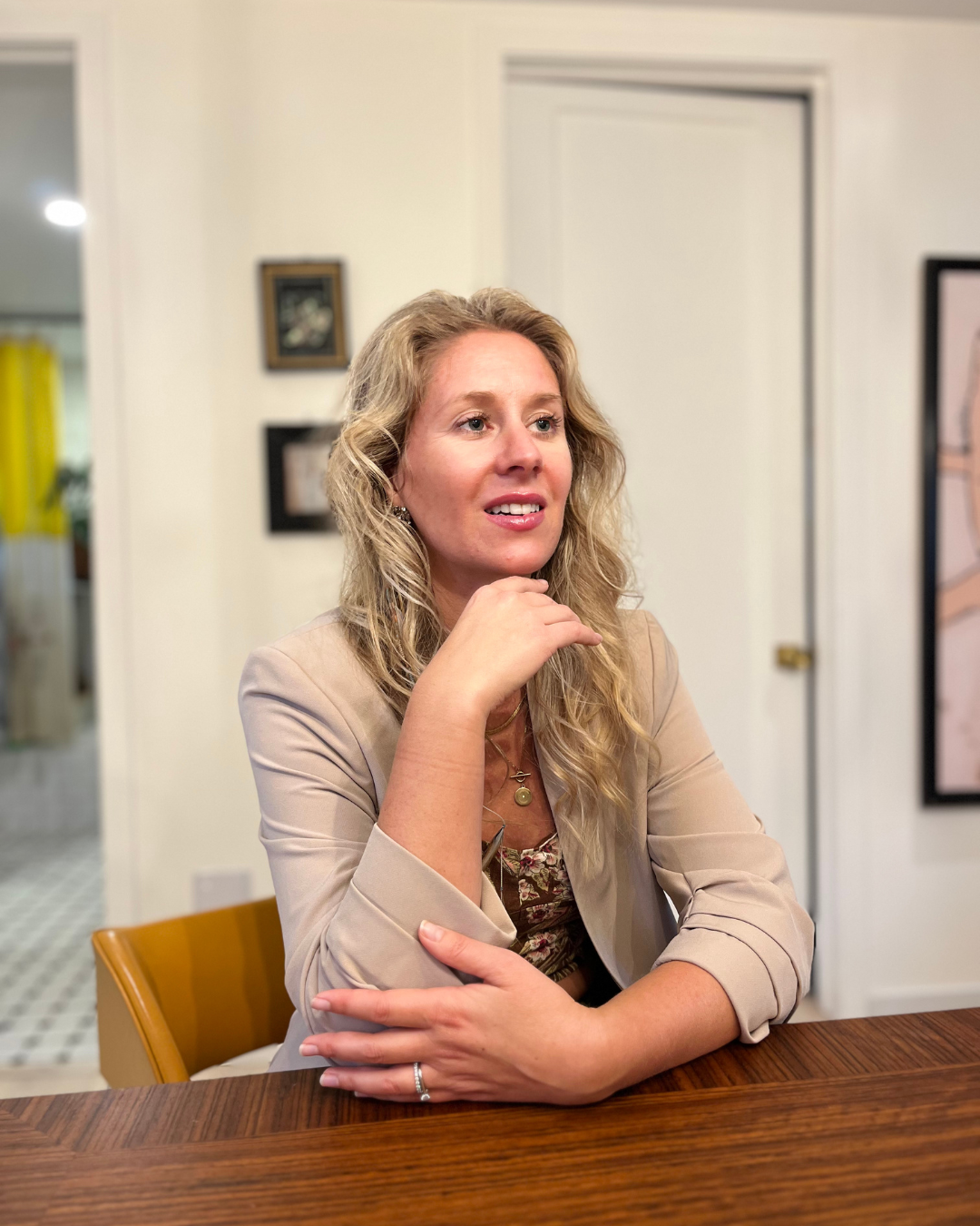 Une femme aux cheveux blonds, habillée d'une veste beige, assise à une table en bois, l'air pensif ou en conversation dans une pièce domestique.