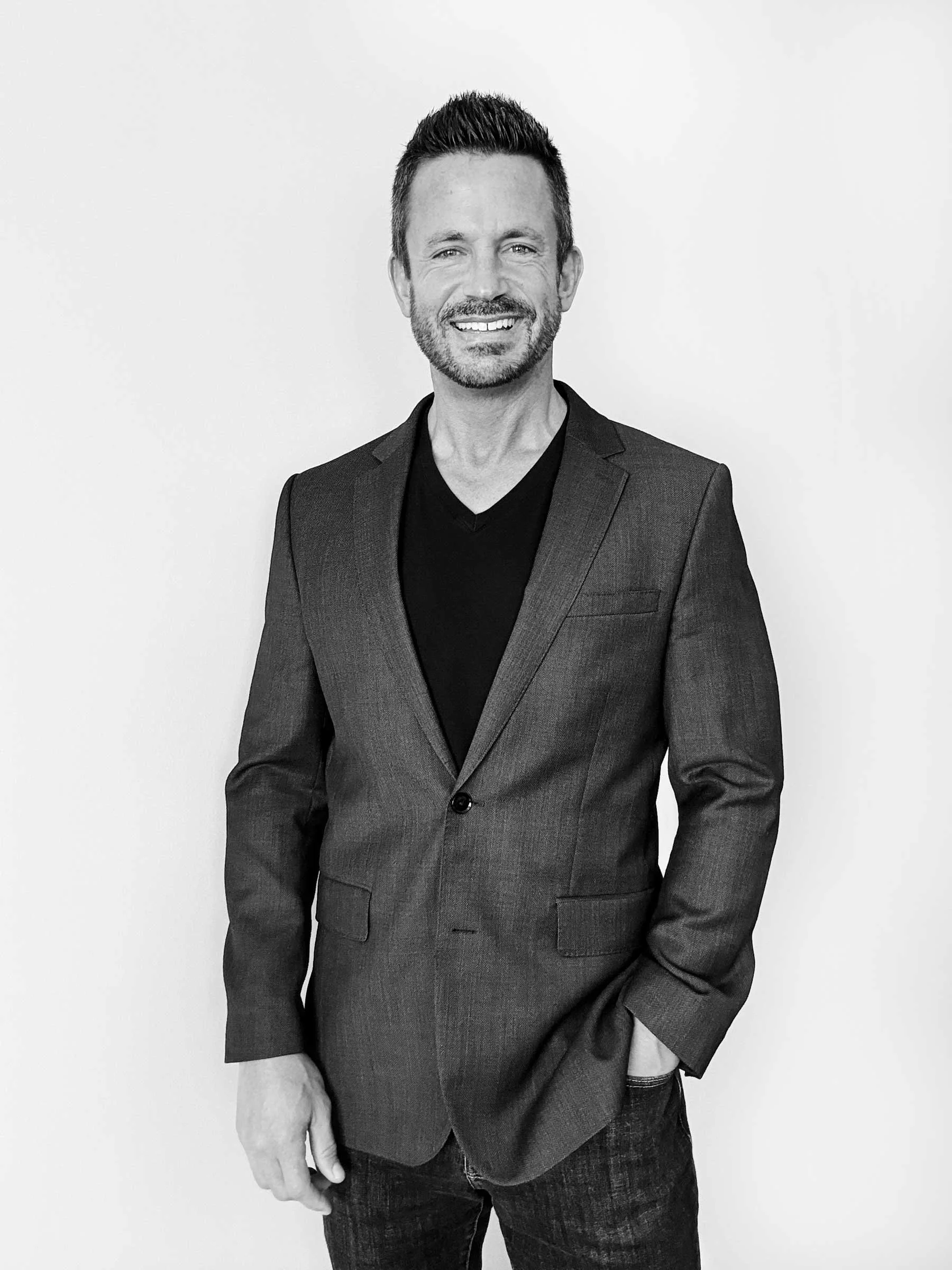 A black and white photo of a smiling man wearing a dark blazer over a dark V-neck shirt, standing against a plain light-colored background.