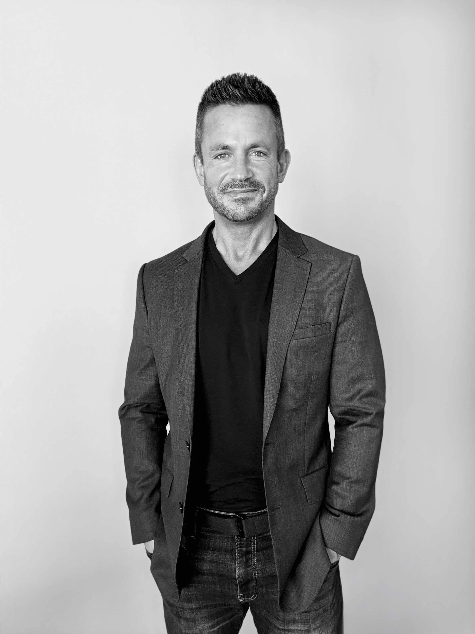Black and white portrait of a man wearing a blazer and jeans, standing with hands in pockets against a plain light background.