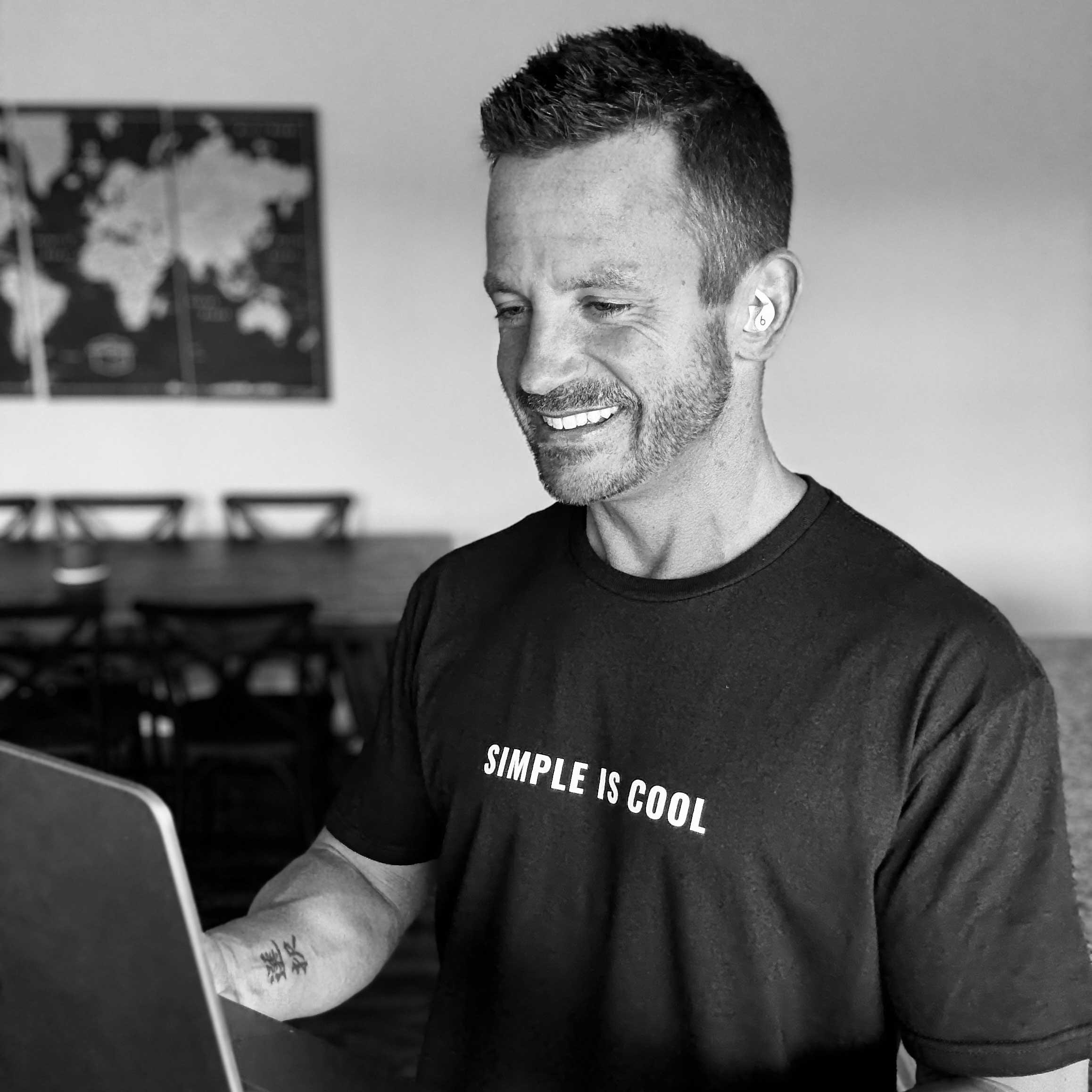 A man smiling and looking at a laptop, wearing a black T-shirt with white text that says 'SIMPLE IS COOL', in a room with chairs and a world map on the wall in the background.