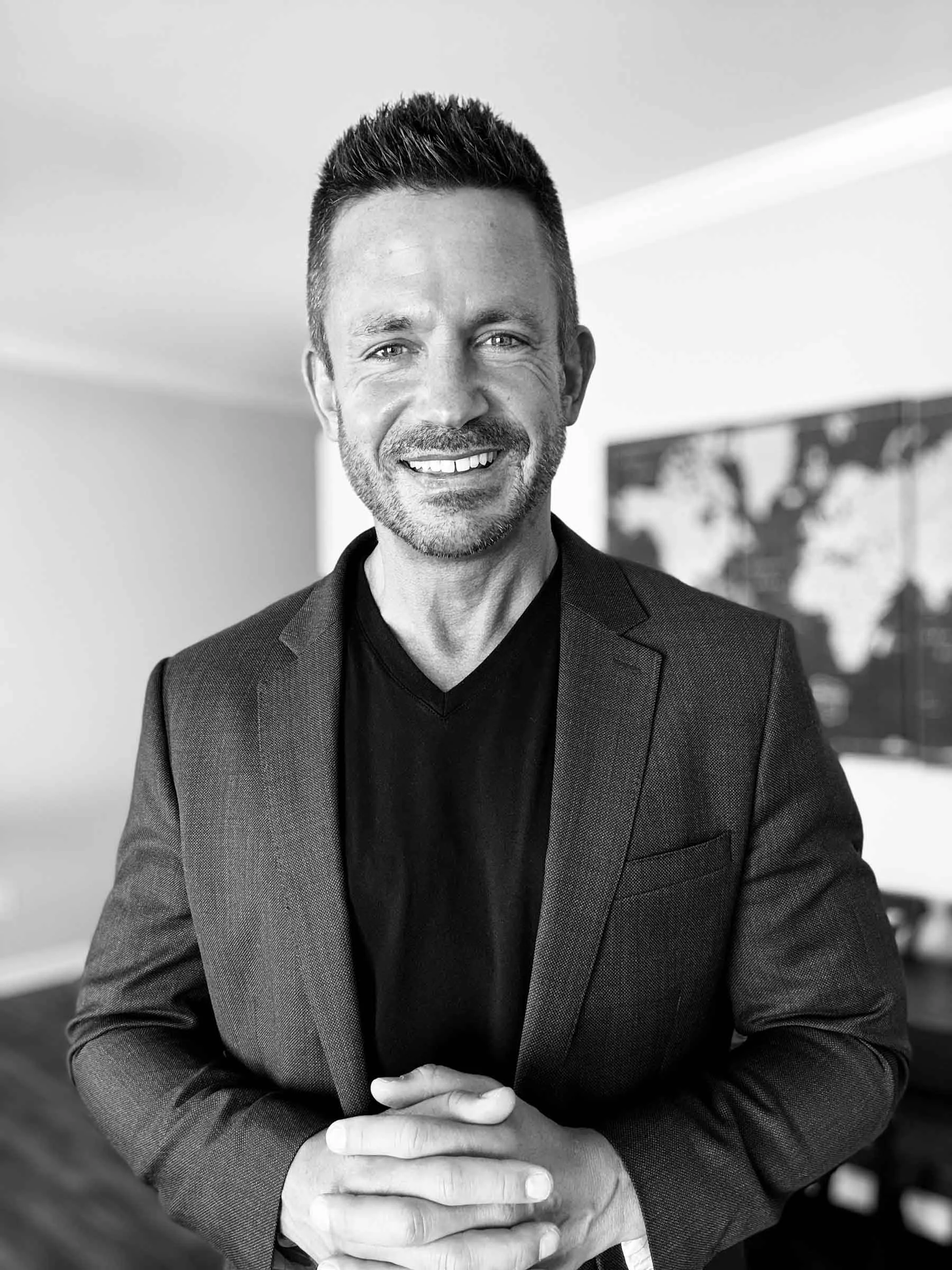 Black and white photo of a man smiling, wearing a suit, with his head slightly tilted, hands clasped, standing indoors with a blurred artwork in the background.
