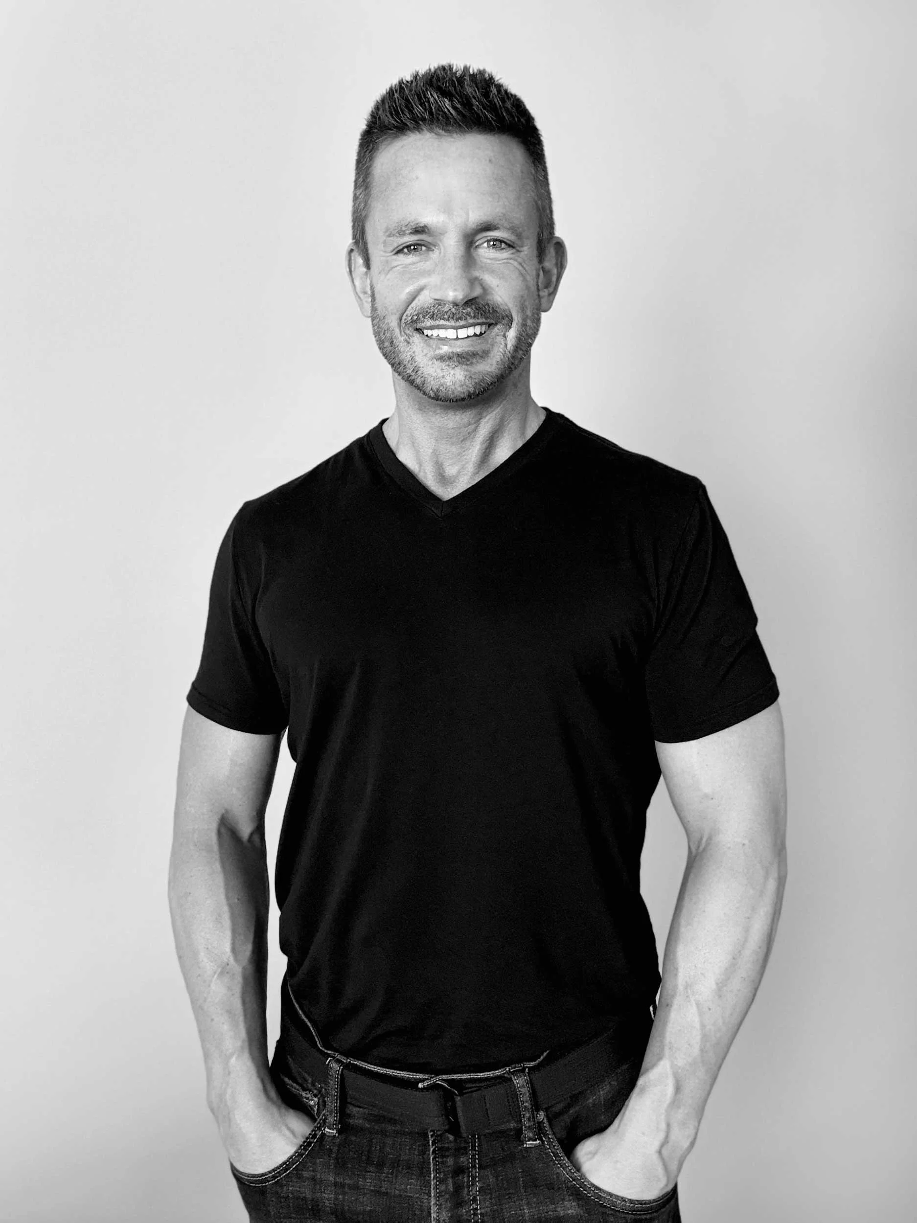 Black and white portrait of a man with short hair and a beard, smiling and wearing a black t-shirt, standing against a plain background.