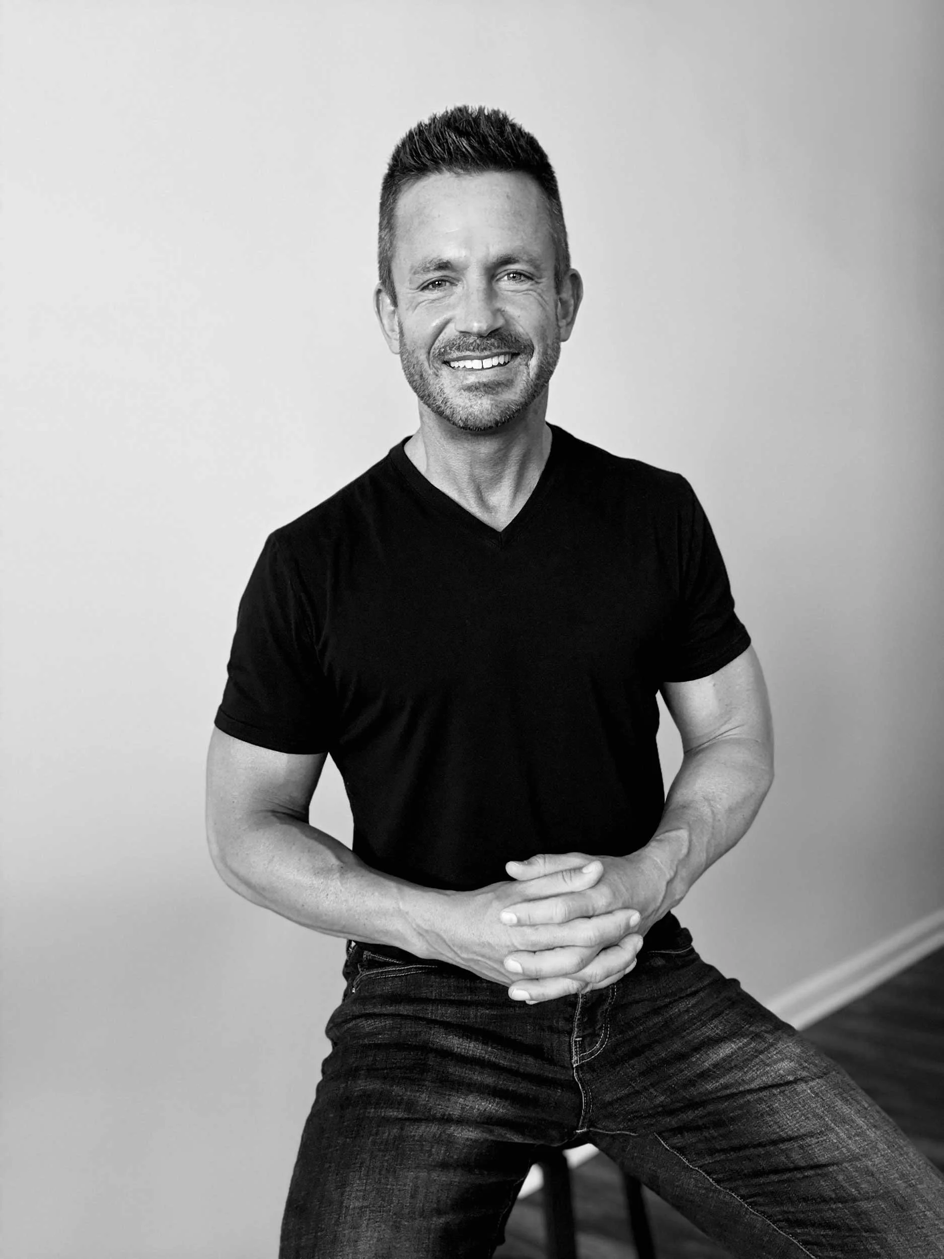 Black-and-white photo of a smiling man with short hair and a beard, wearing a black t-shirt, standing against a plain wall.