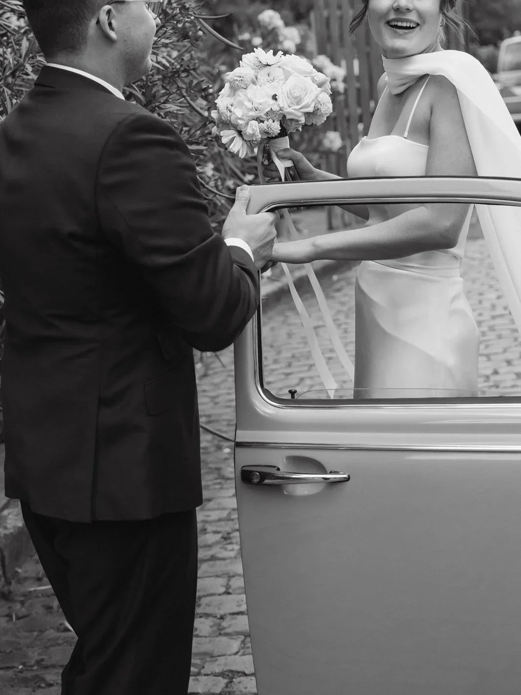 Ein Mann in einem schwarzen Anzug übergibt einer lachenden Frau im Hochzeitskleid einen Blumenstrauß beim Verlassen eines Autos.