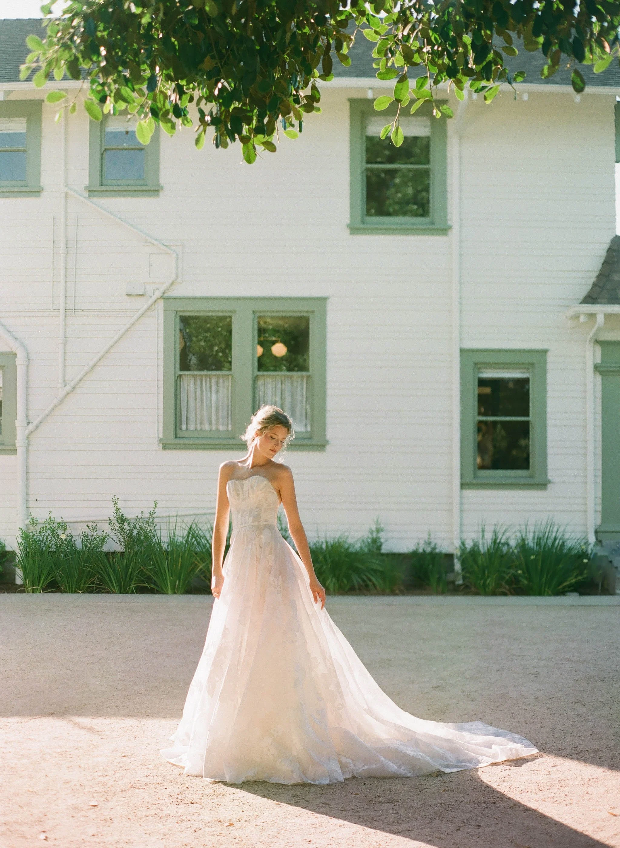 San Juan Capistrano, A young woman in a strapless white wedding dress standing outdoors on a sandy surface, with a white house and green plants behind her, and sunlight casting a shadow.