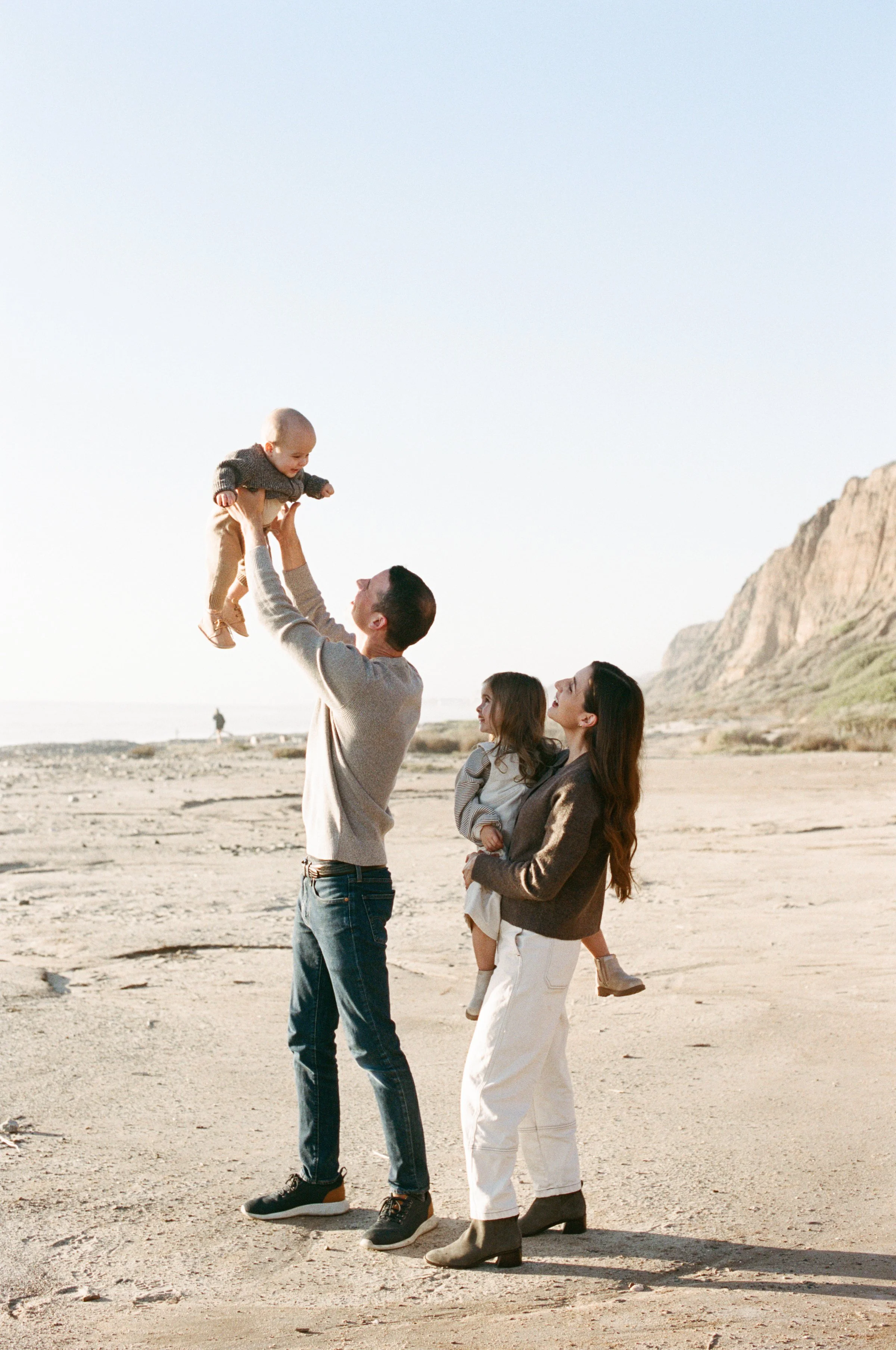 A family of four enjoying a day at the beach, with father lifting a baby into the air, mother holding a young girl, and scenic cliffs in the background.