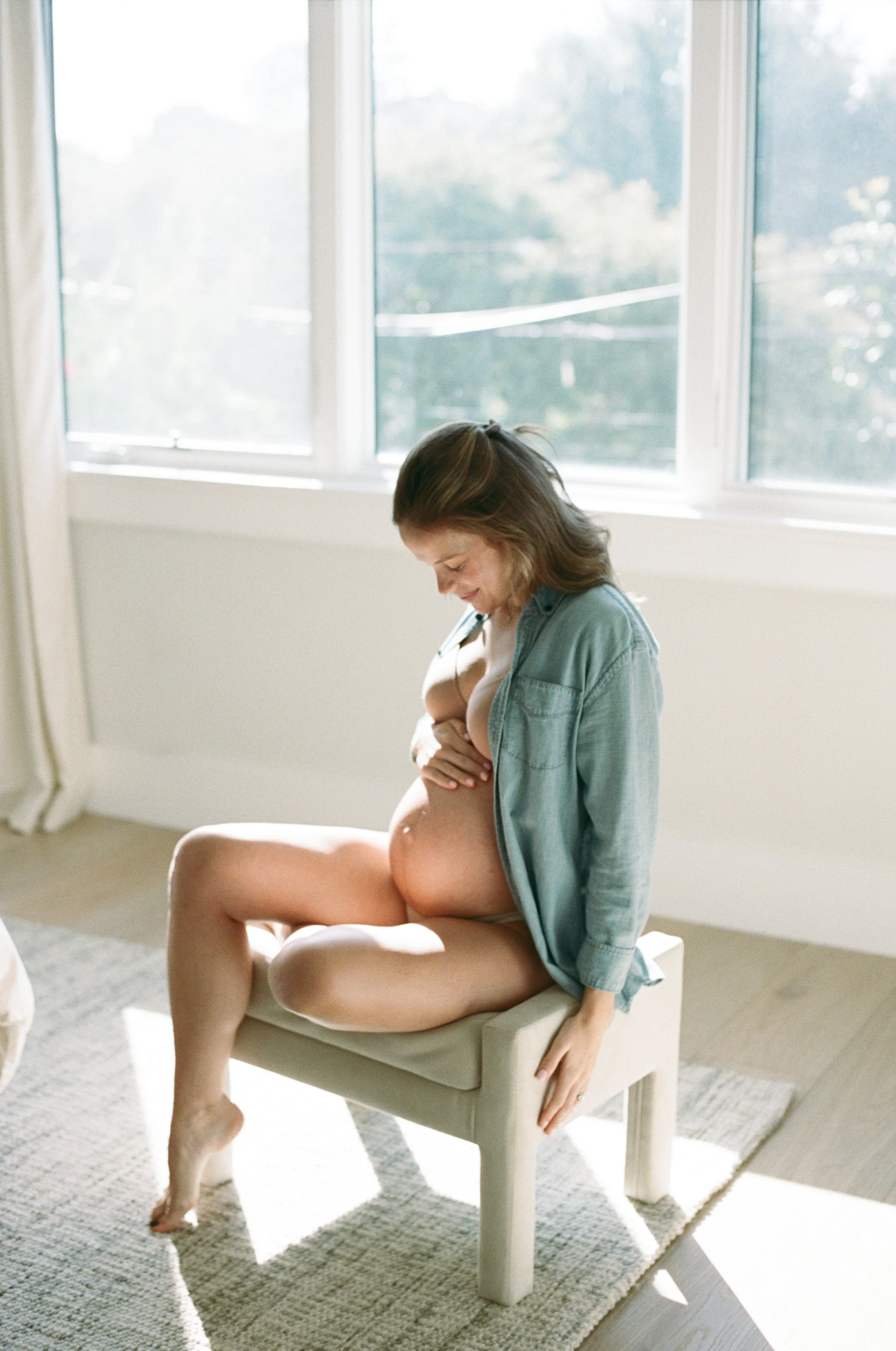 photography of a pregnant woman sitting on a bench near a window, smiling and cradling her belly San Clemente, Orange County