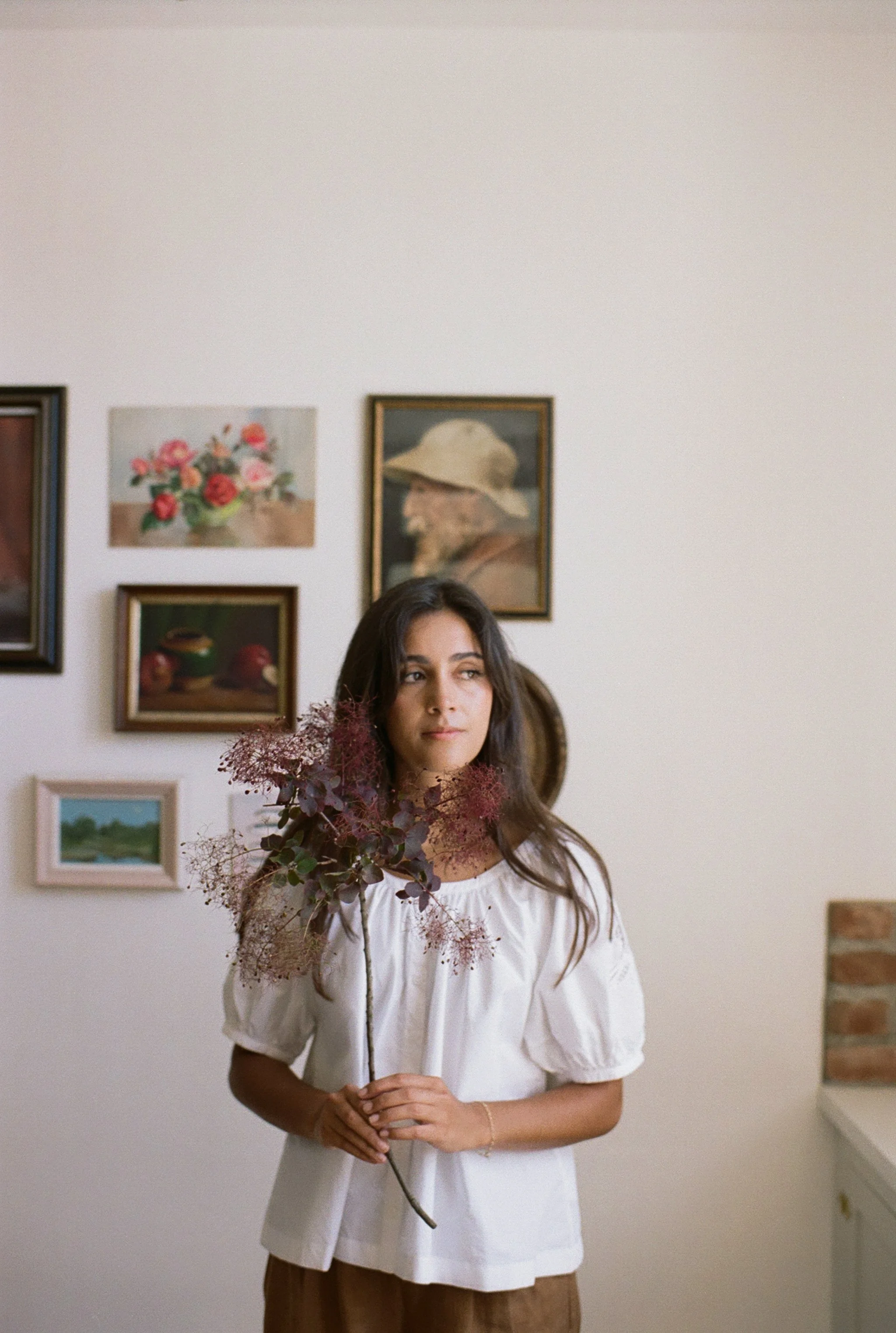 A woman with long dark hair in a white blouse holding a bouquet with purple and pink flowers inside, standing in a room with framed artwork on the wall.