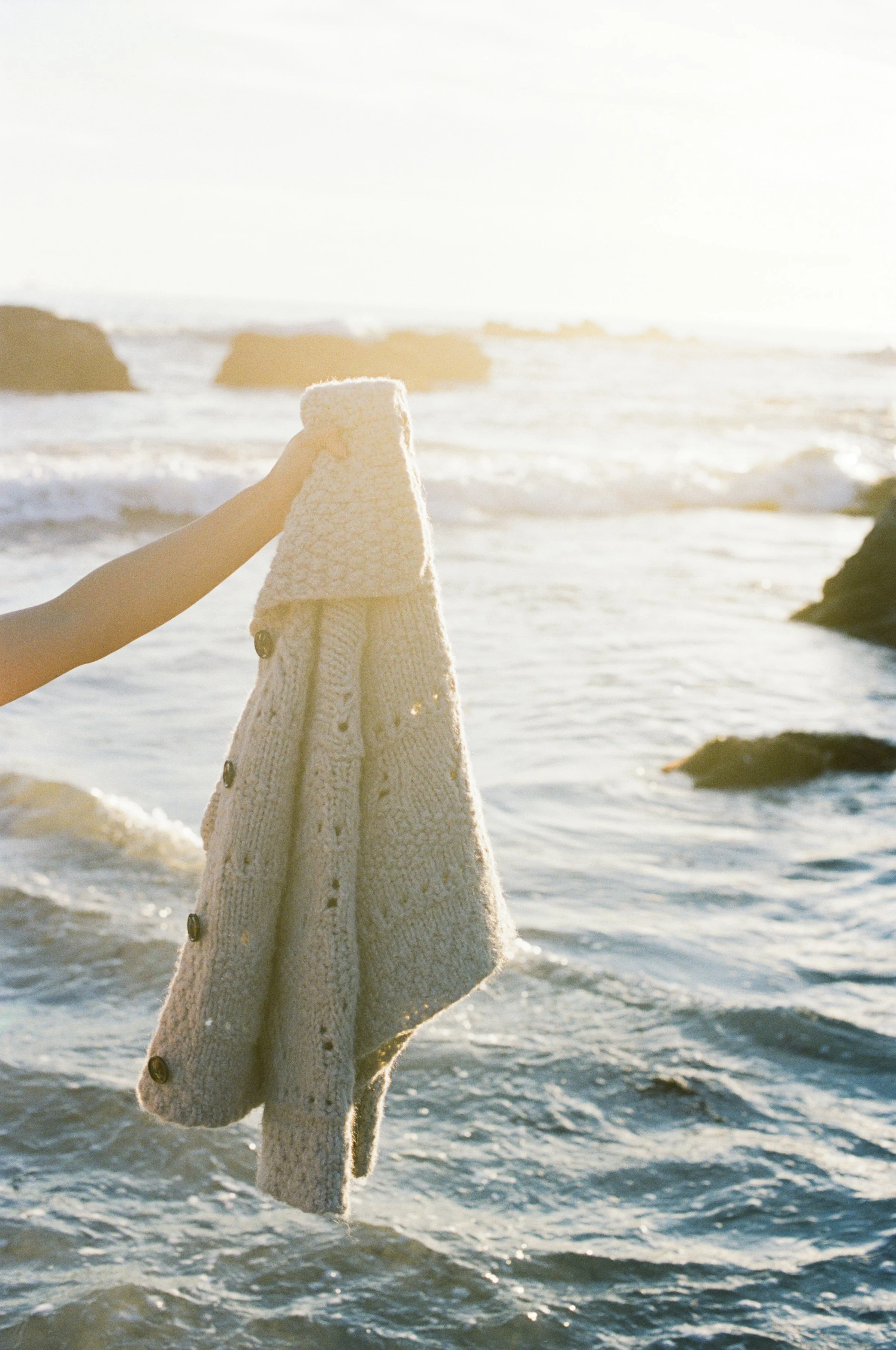 Person holding a beige knitted sweater over a body of water at the beach during sunset.