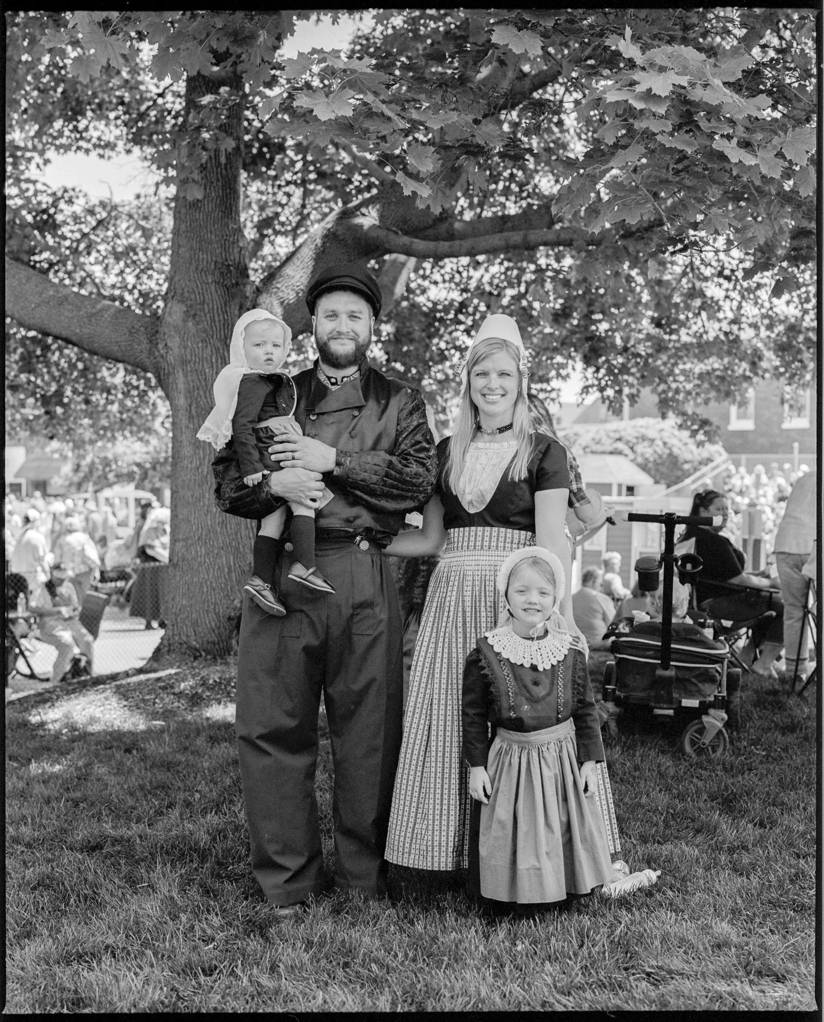 A family dressed in traditional or historical costumes standing on grass under a large tree, with a crowd in the background suggesting an outdoor event or festival. Pella, Iowa Tulip Time Festival, Dutch Heritage