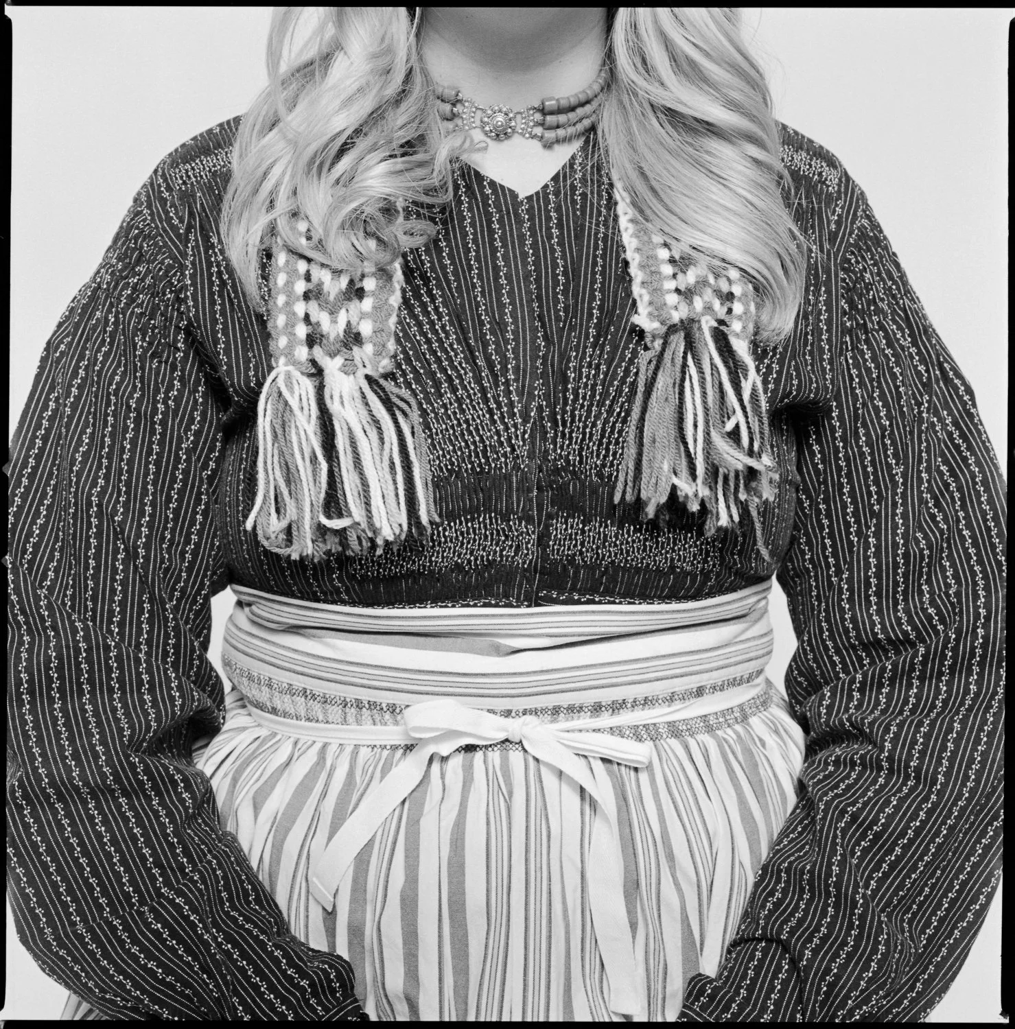 Black and white photo of a woman from the neck down, wearing a patterned blouse with embroidery, a beaded necklace, and an apron with a striped pattern tied with a bow. Pella, Iowa Tulip Time Festival, Dutch Heritage