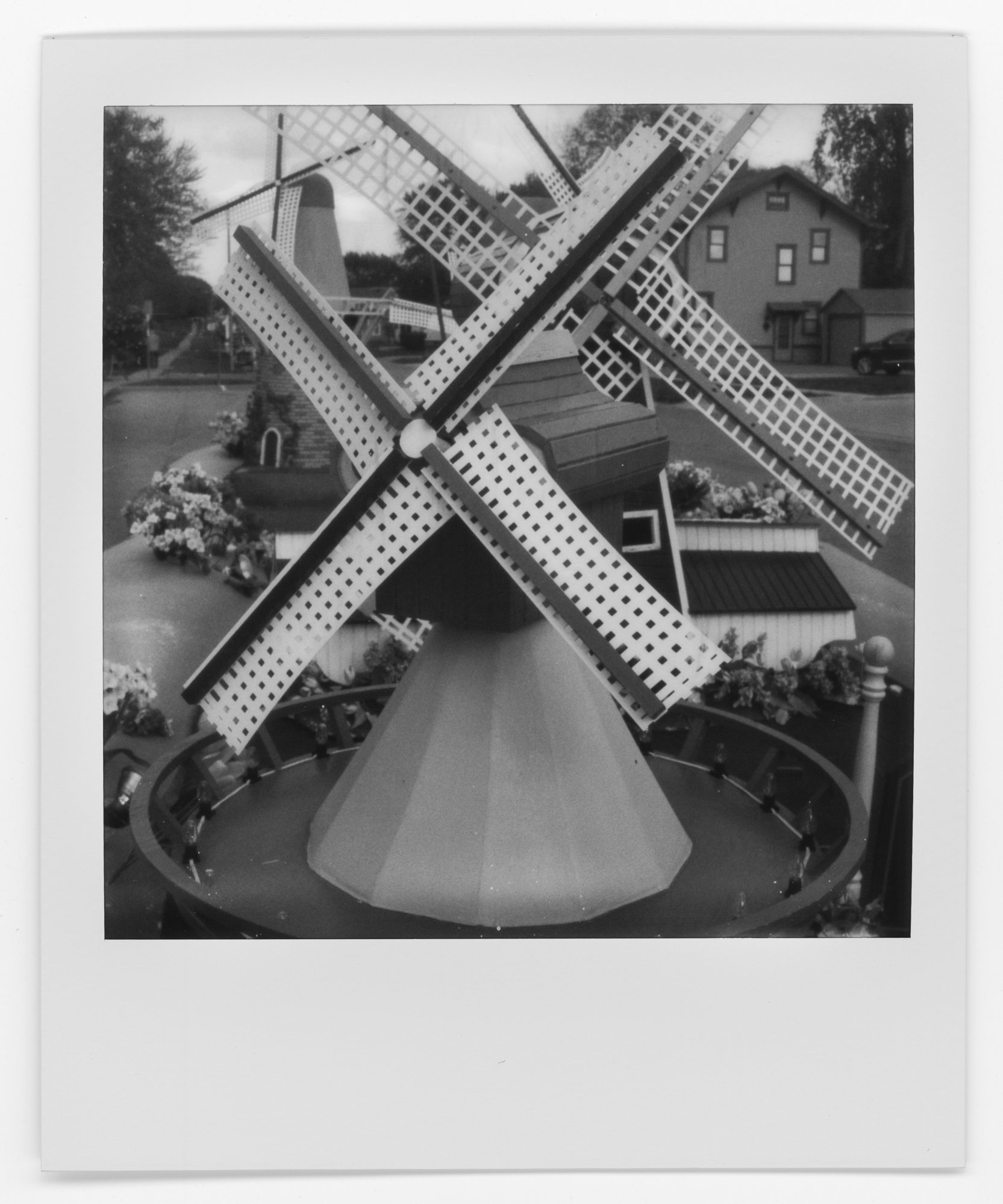 Black and white photo of a small windmill with four blades, surrounded by flowers and small fence, located outdoors in a residential neighborhood. Pella, Iowa Tulip Time Festival, Dutch Heritage