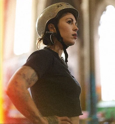A woman wearing a motorcycle helmet and a black T-shirt, looking to the side with arm tattoos visible. Sk8 Liborius