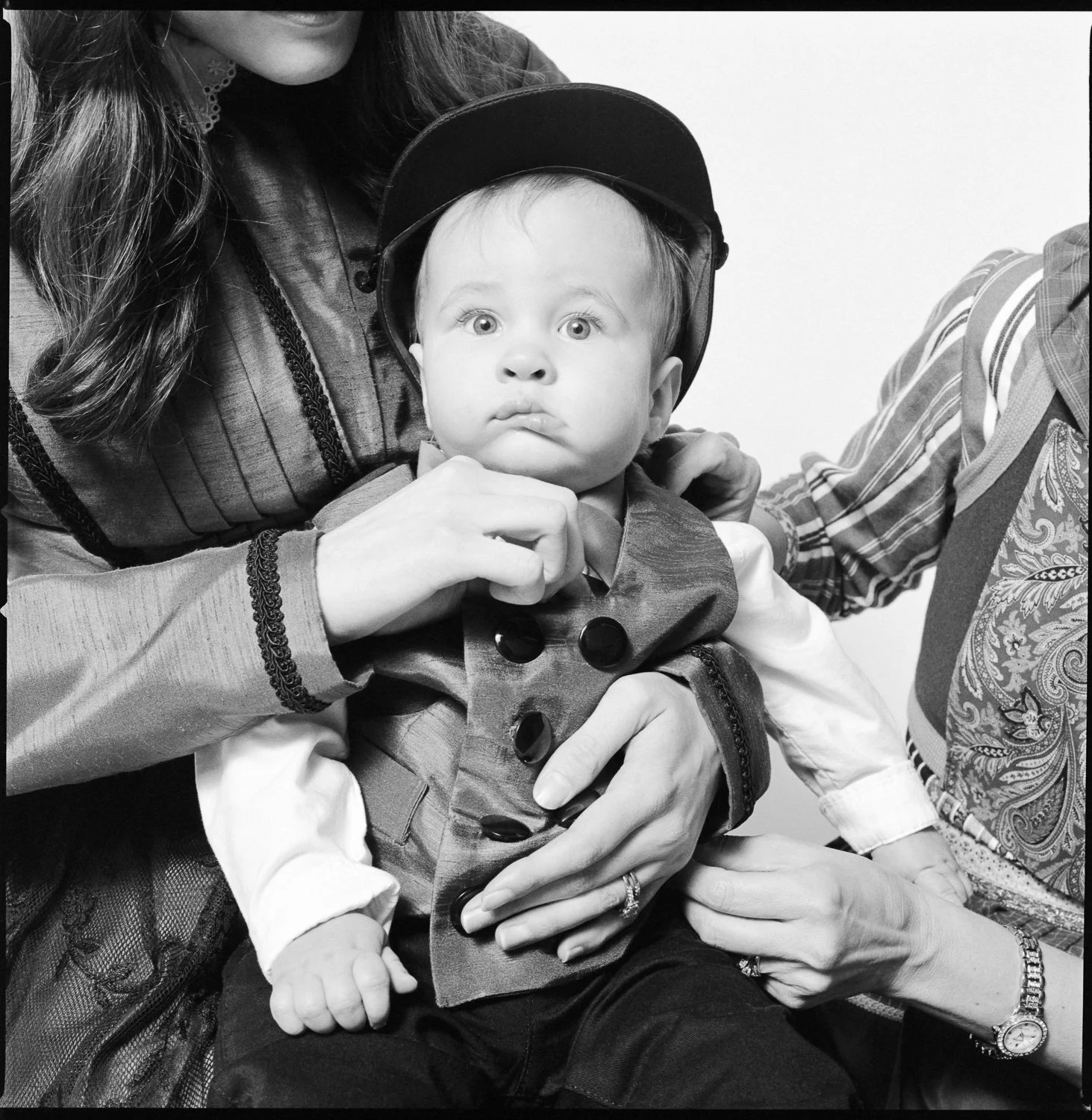 A young child with wide eyes and a serious expression, wearing a cap and a fancy vest, being held by two adults, one on each arm. The image is in black and white. Pella, Iowa Tulip Time Festival, Dutch Heritage