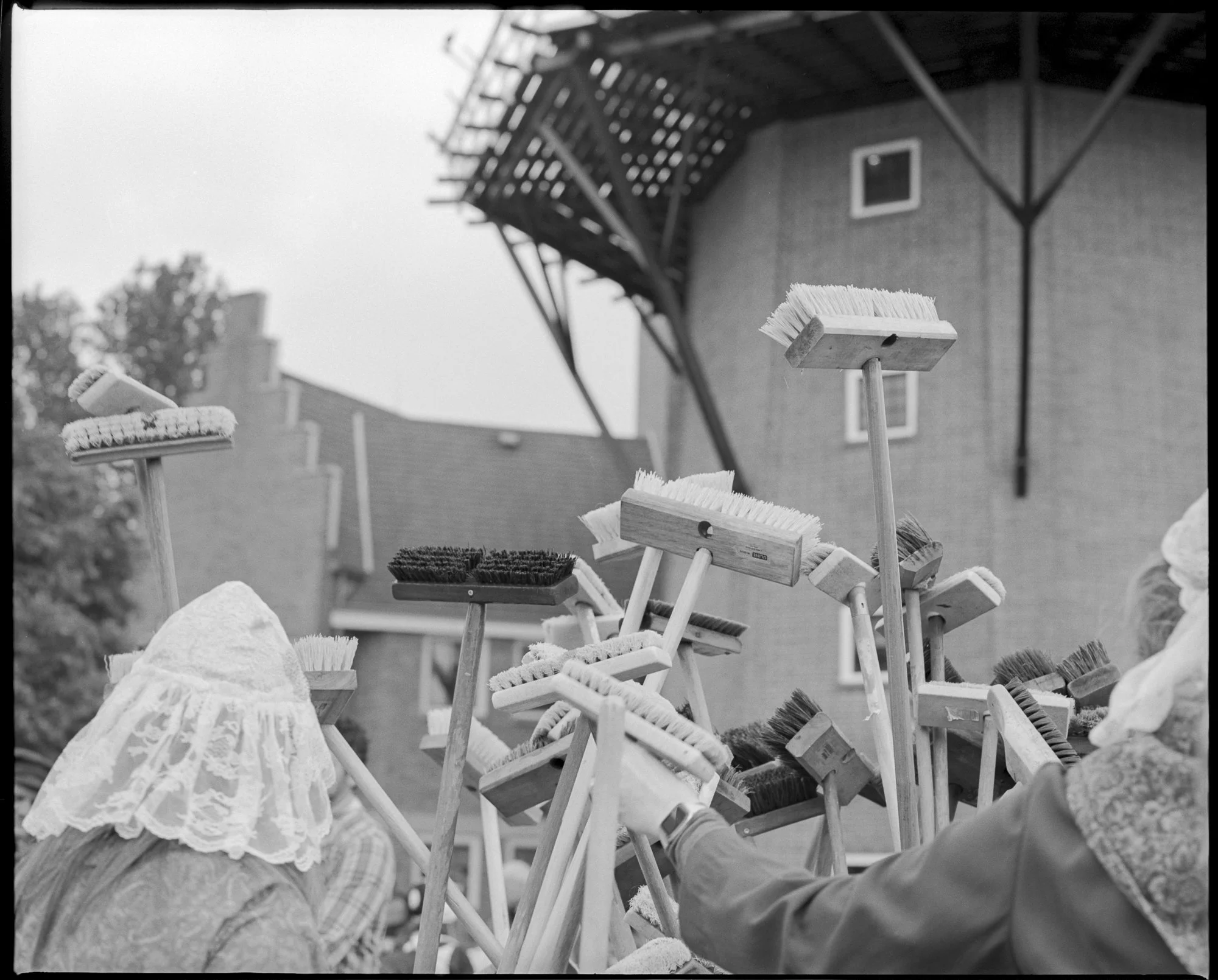 A collection of brooms with various handle lengths and bristle styles, held upright and arranged outdoors near a brick building with a staircase in the background. Pella, Iowa Tulip Time Festival, Dutch Heritage