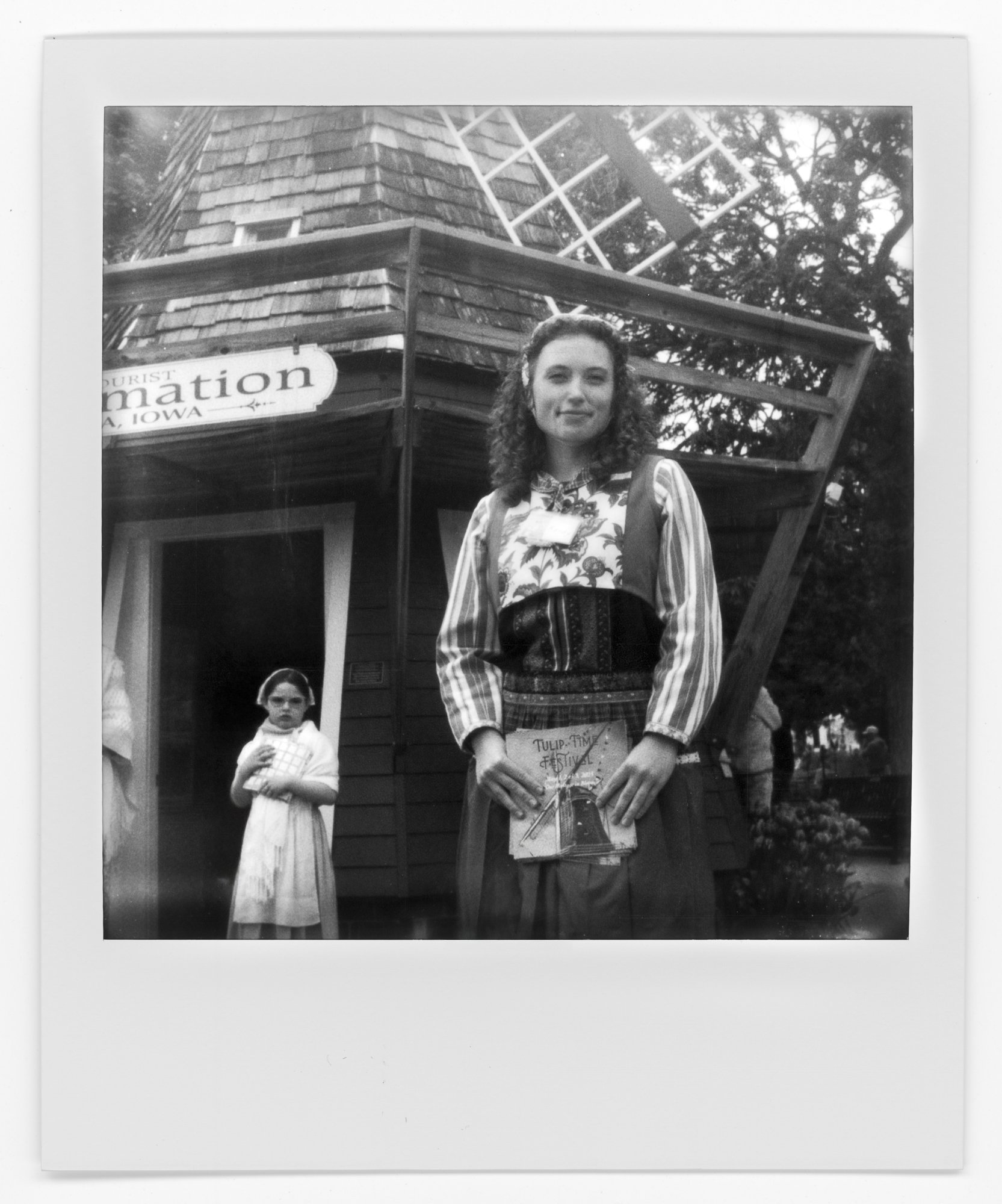 A young woman in traditional clothing holding a booklet, standing in front of a wooden building with a sign that reads 'Tourist Information' in Iowa, with a girl in a dress in the background and trees around. Pella, Iowa Tulip Time Festival, Dutch He