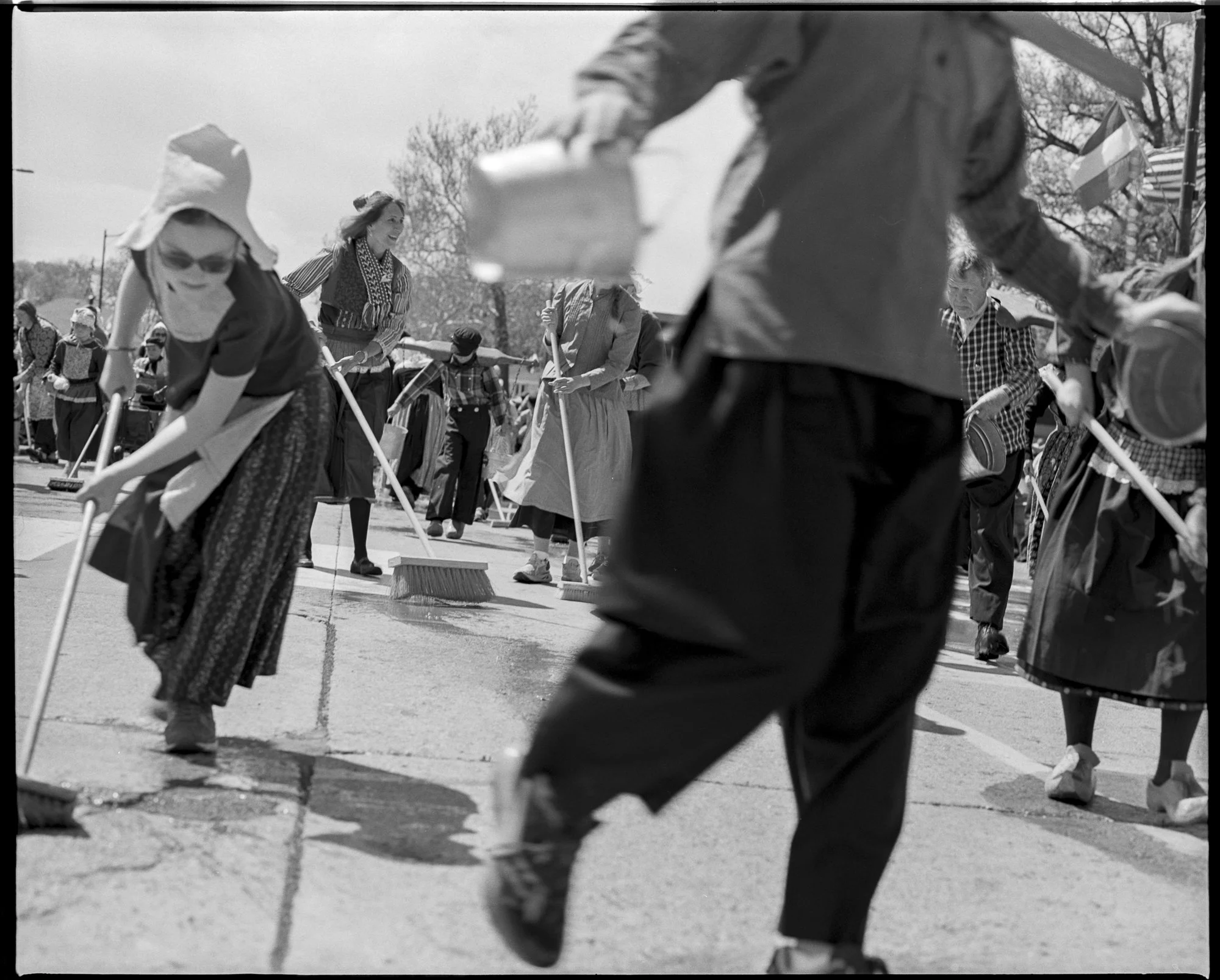 People cleaning and sweeping outdoor pavement during a community cleanup event. Some participants are wearing hats and casual clothing. Pella, Iowa Tulip Time Festival, Dutch Heritage