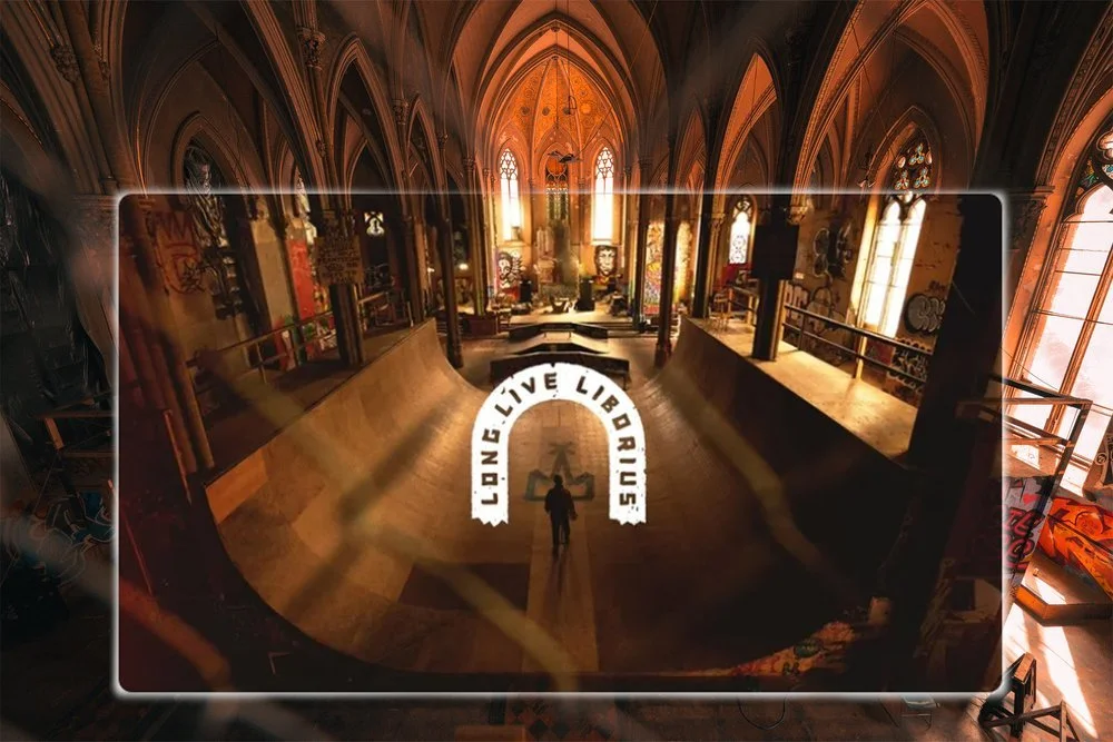 Interior of a Gothic-style church or cathedral with tall arched windows and high vaulted ceilings. There is graffiti on the walls, a sponsored skateboarding half-pipe in the center, and a person standing on the half-pipe with a sign that reads "LONG LIVE LIBRULIS."