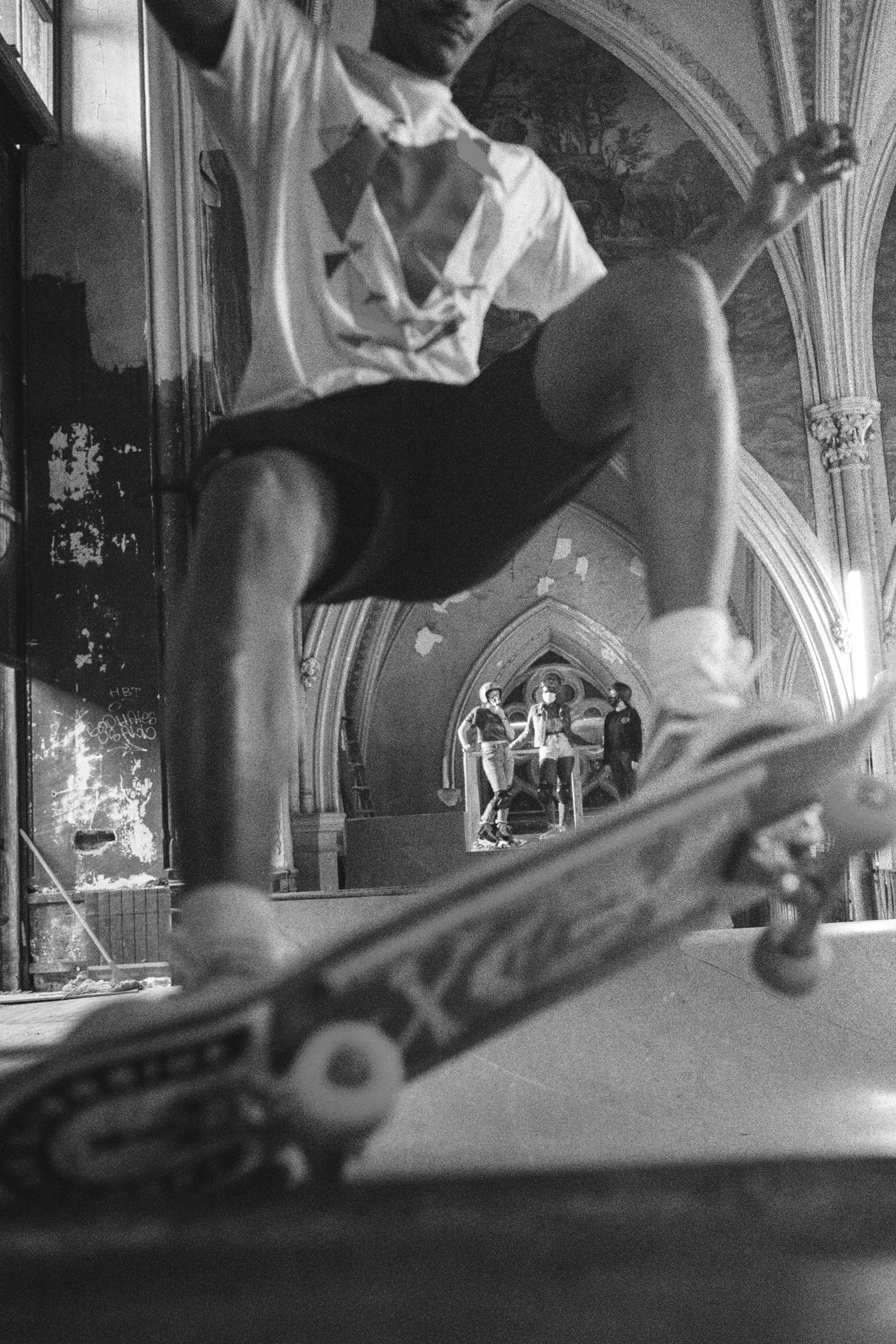 A person skateboarding indoors with a group of people standing on a raised platform in the background, in a building with high, ornate ceilings. Sk8 Liborius